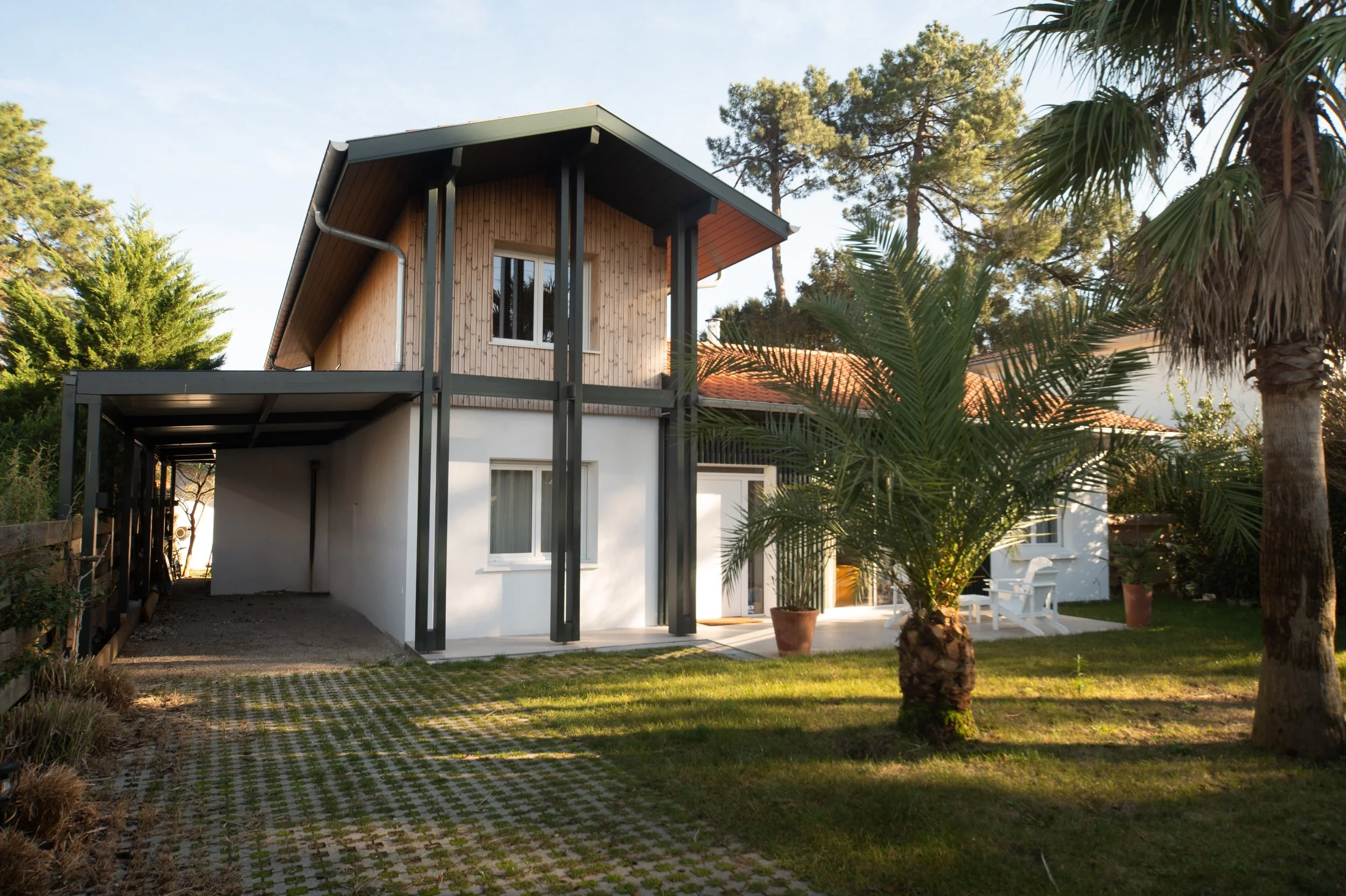 Maison moderne avec deux étages, façade rez de chaussée blanche et étage supérieur en bardage bois, toit en tuiles rouges, jardin avec palmiers, espace de détente et allée pavée.