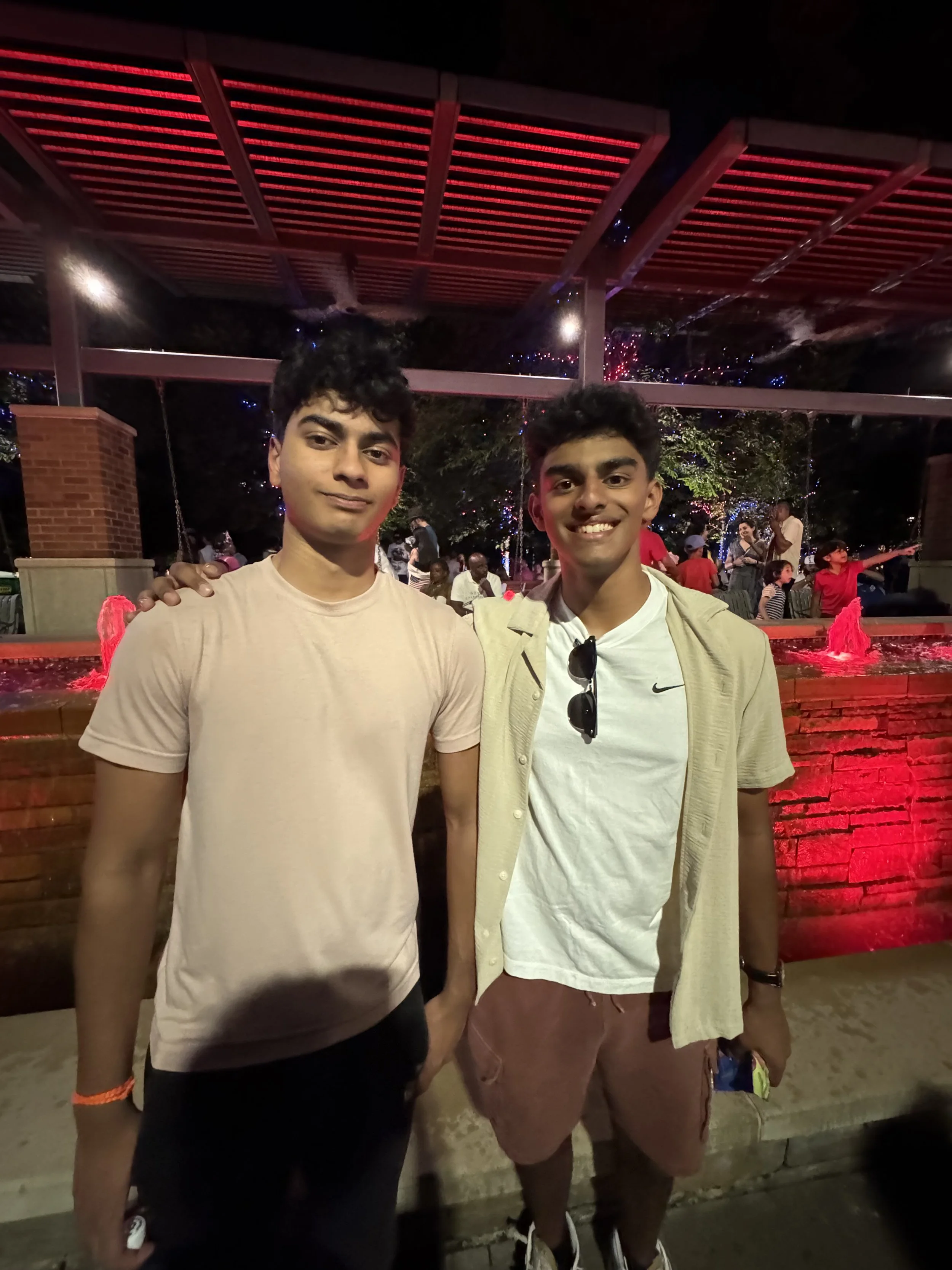 Two young men standing together at an outdoor night event near a fountain with red lighting, surrounded by people and festive decorations.