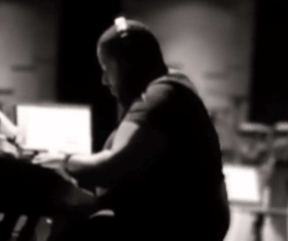 A woman with short hair sitting at a desk working on a computer in a dimly lit room.