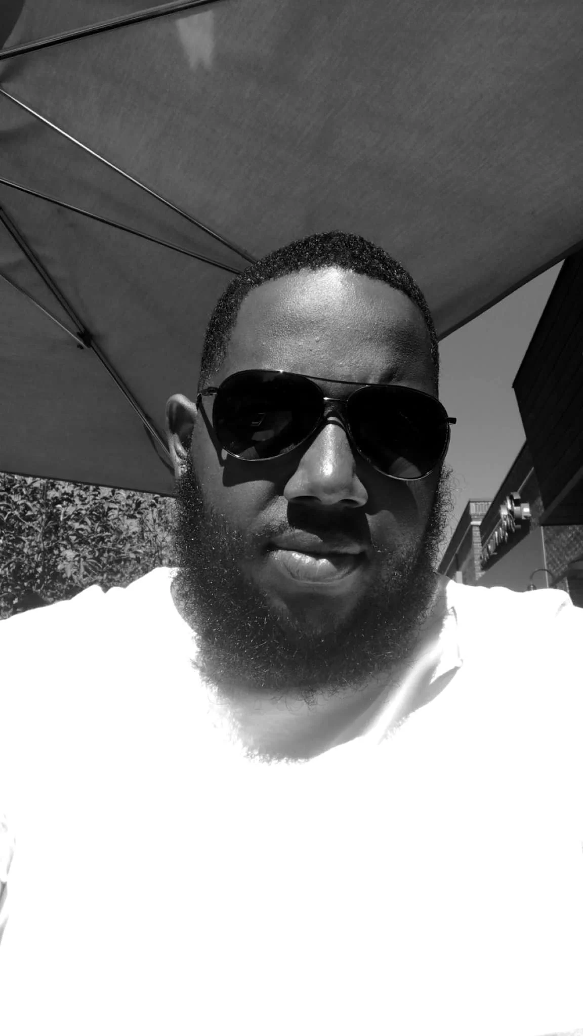 A man wearing dark sunglasses and a white shirt taking a selfie outdoors on a sunny day.