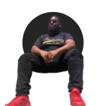 A man sitting against a dark background, wearing a dark t-shirt and pants with red shoes.