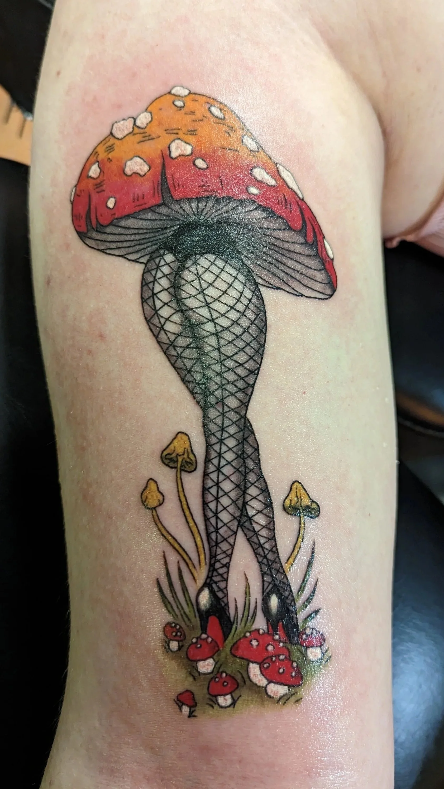 Tattoo of a large mushroom with a red cap, black and white stem, and fishnet stockings, surrounded by smaller mushrooms and grass.