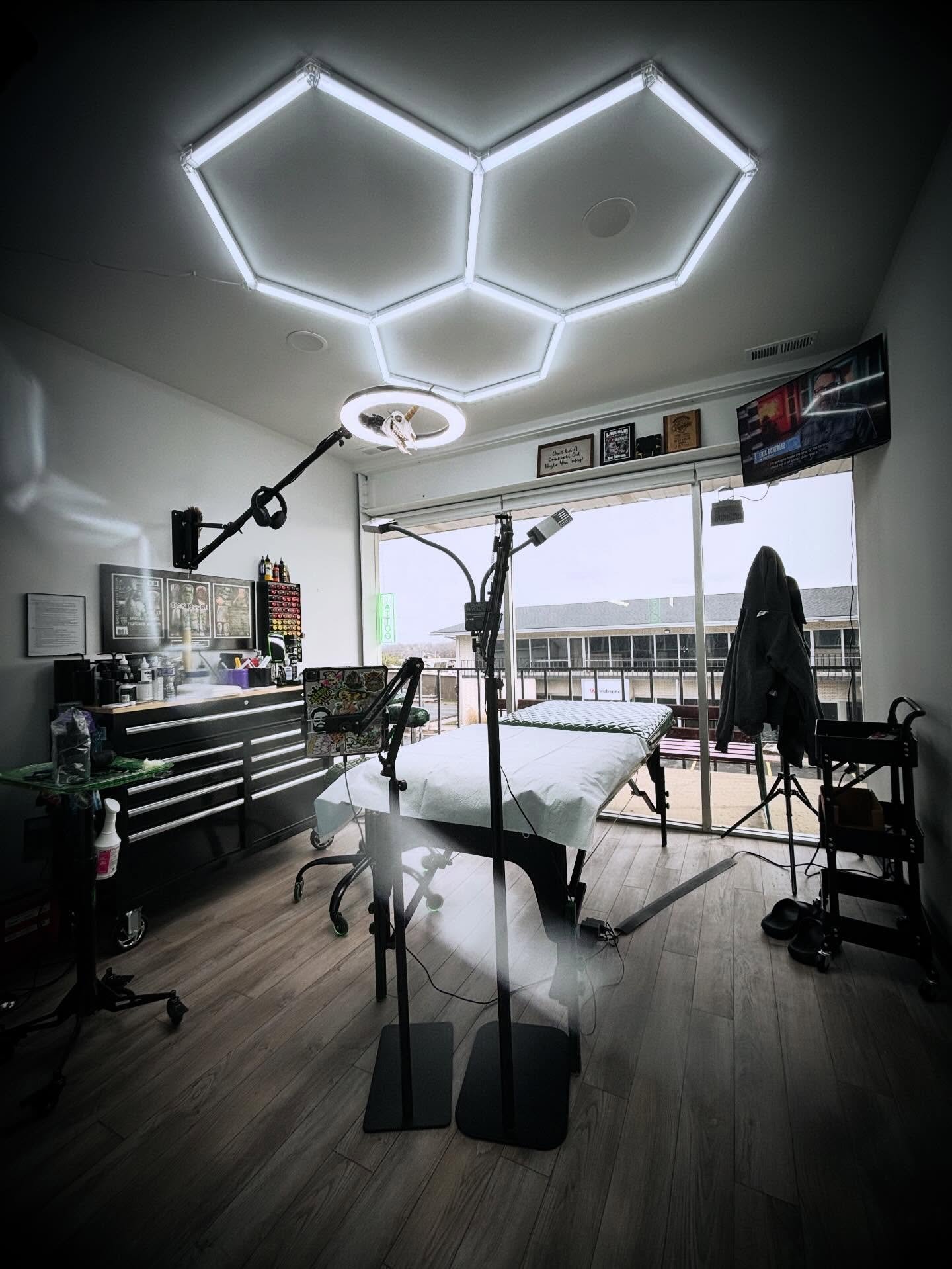 A tattoo studio with a massage table, overhead hexagonal LED lights, and tattoo equipment, including a tattoo machine and supplies, near large windows.