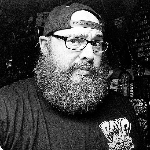 A man with a beard and glasses wearing a black baseball cap backward, standing in a store or workshop with various items in the background.