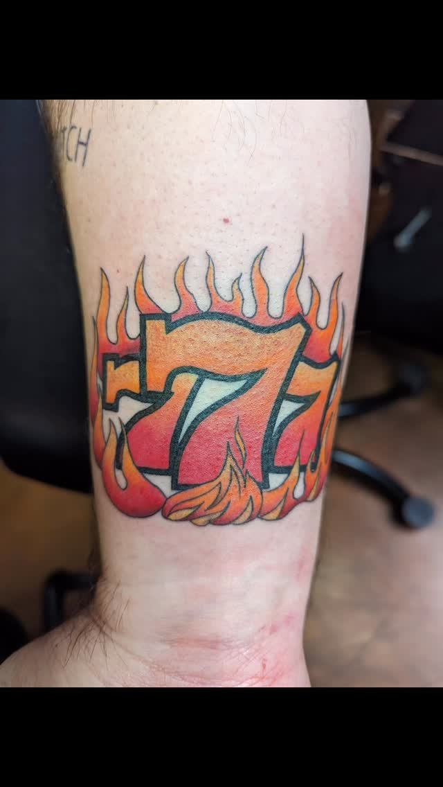 Tattoo of the number 7 surrounded by flames, with orange, red, and yellow colors.