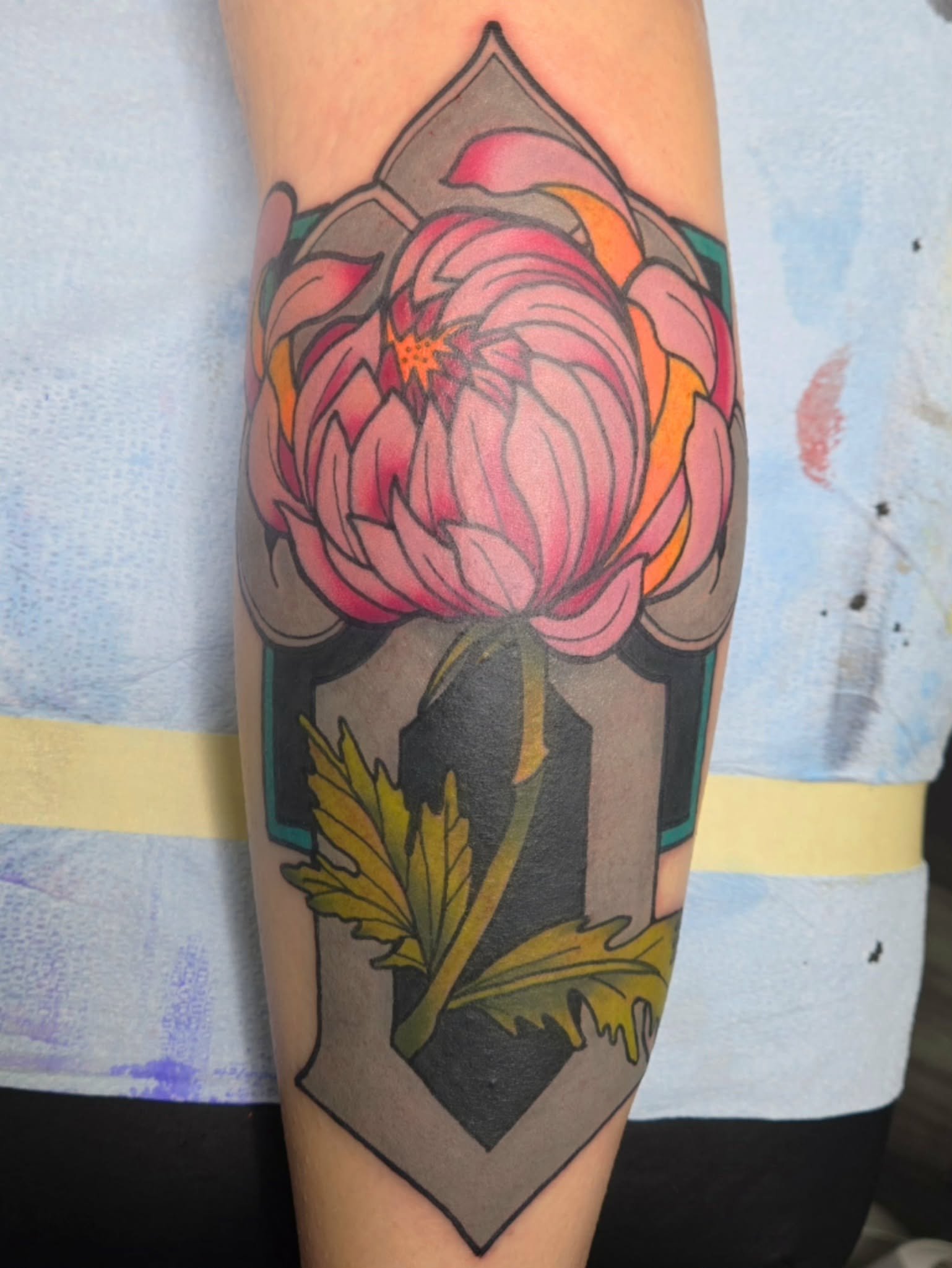 Tattoo of a pink peony flower with green leaves inside a decorative black and gray diamond-shaped frame on person's arm.