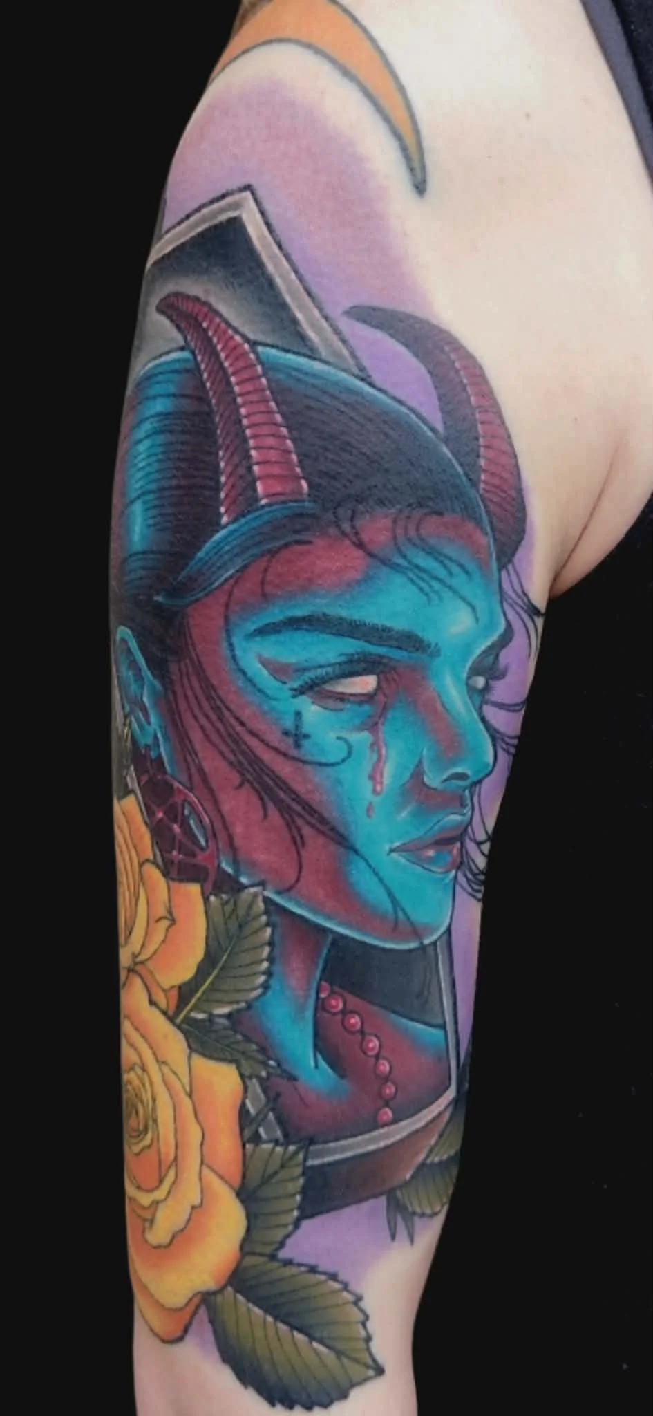 Colorful tattoo depicting a woman's face with blue and purple hues, with horns on her head and tears on her cheeks, accompanied by yellow roses with green leaves.