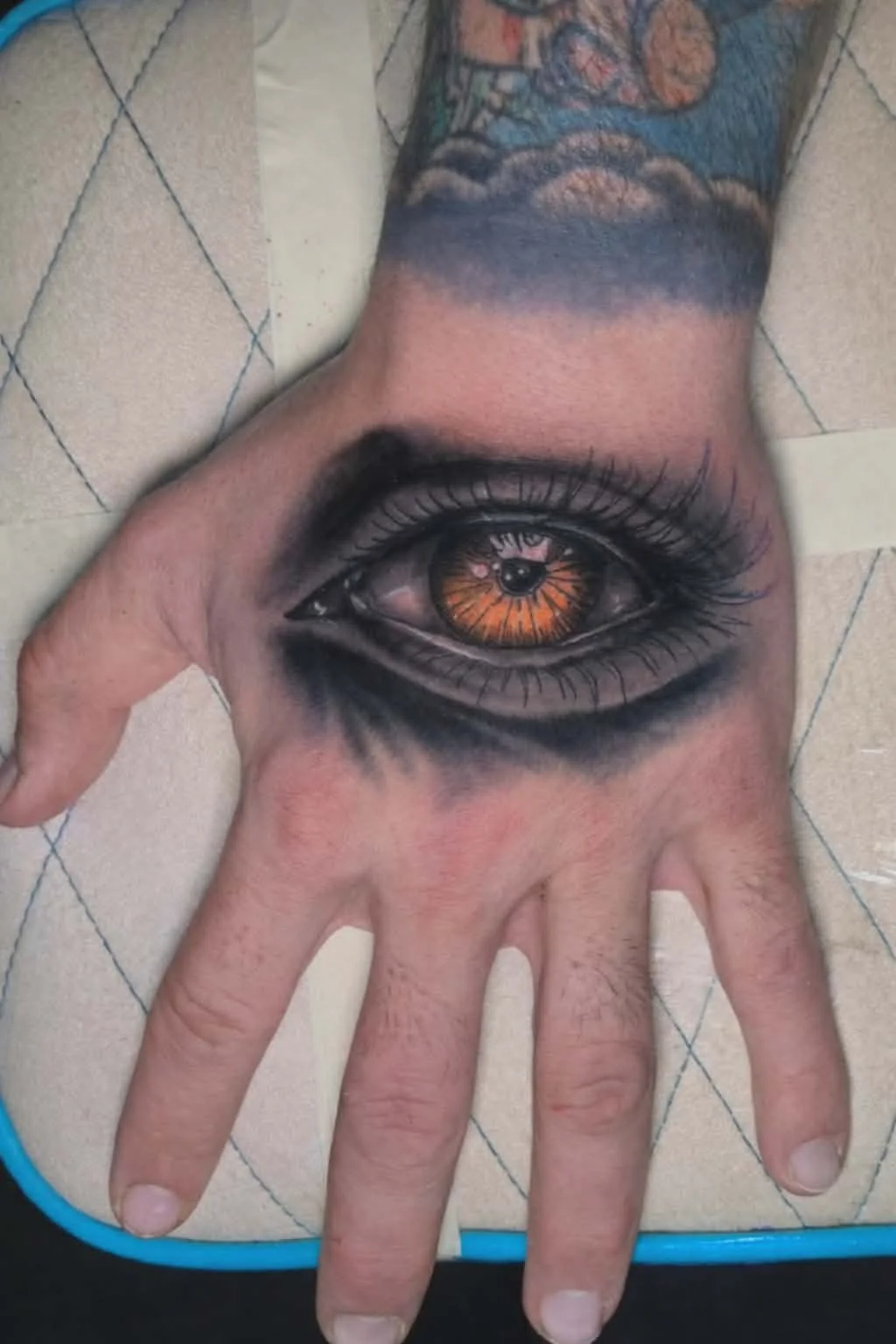 Tattoo of a realistic human eye on a person's hand, with detailed iris and eyelashes, extending onto the wrist and forearm.