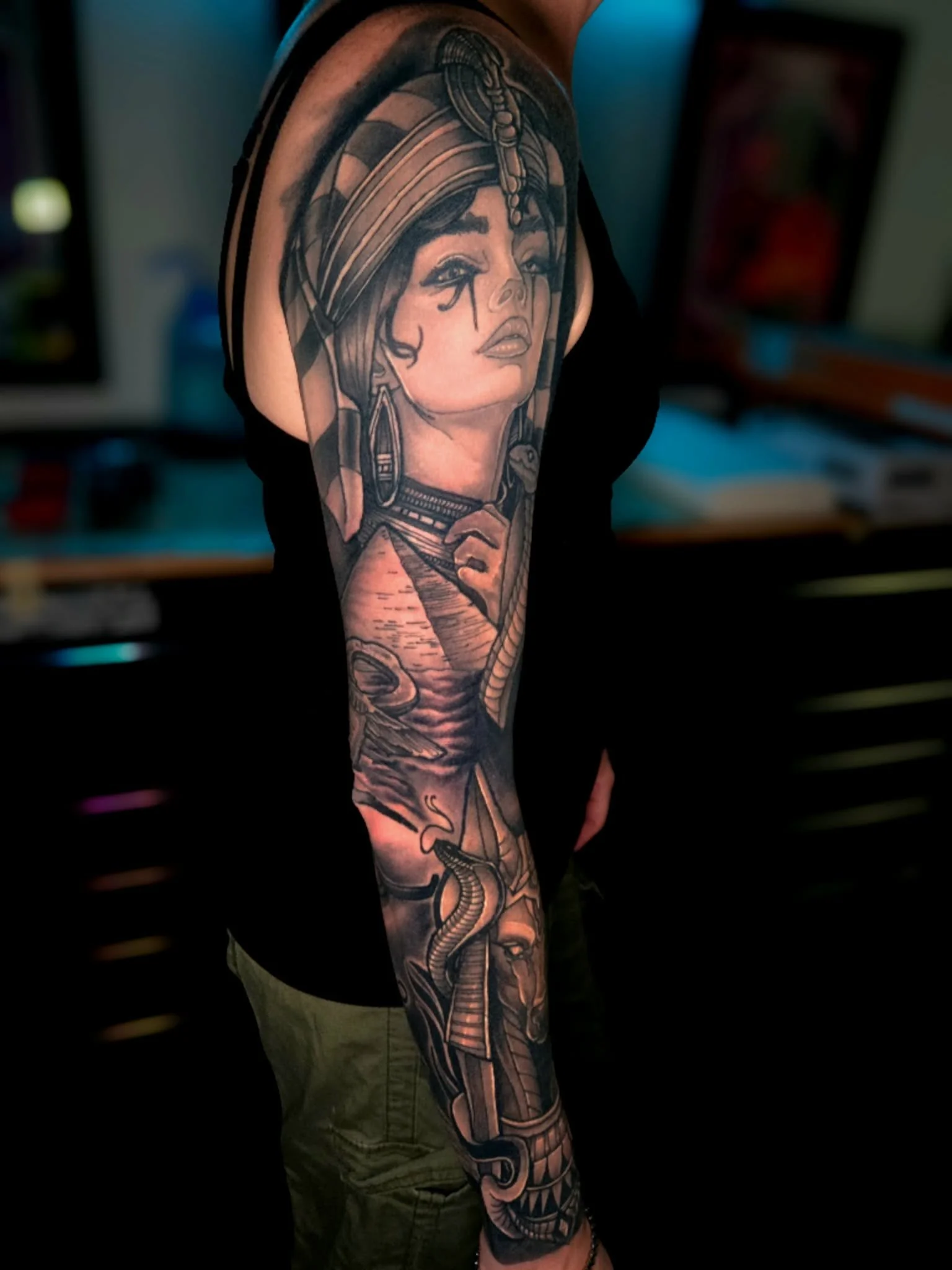 A black and gray tattoo of a woman of indigenous appearance wearing traditional headdress with feathers and beads, earrings, and face paint, horror-themed elements with snakes, a volcano, and a fierce face on the arm.