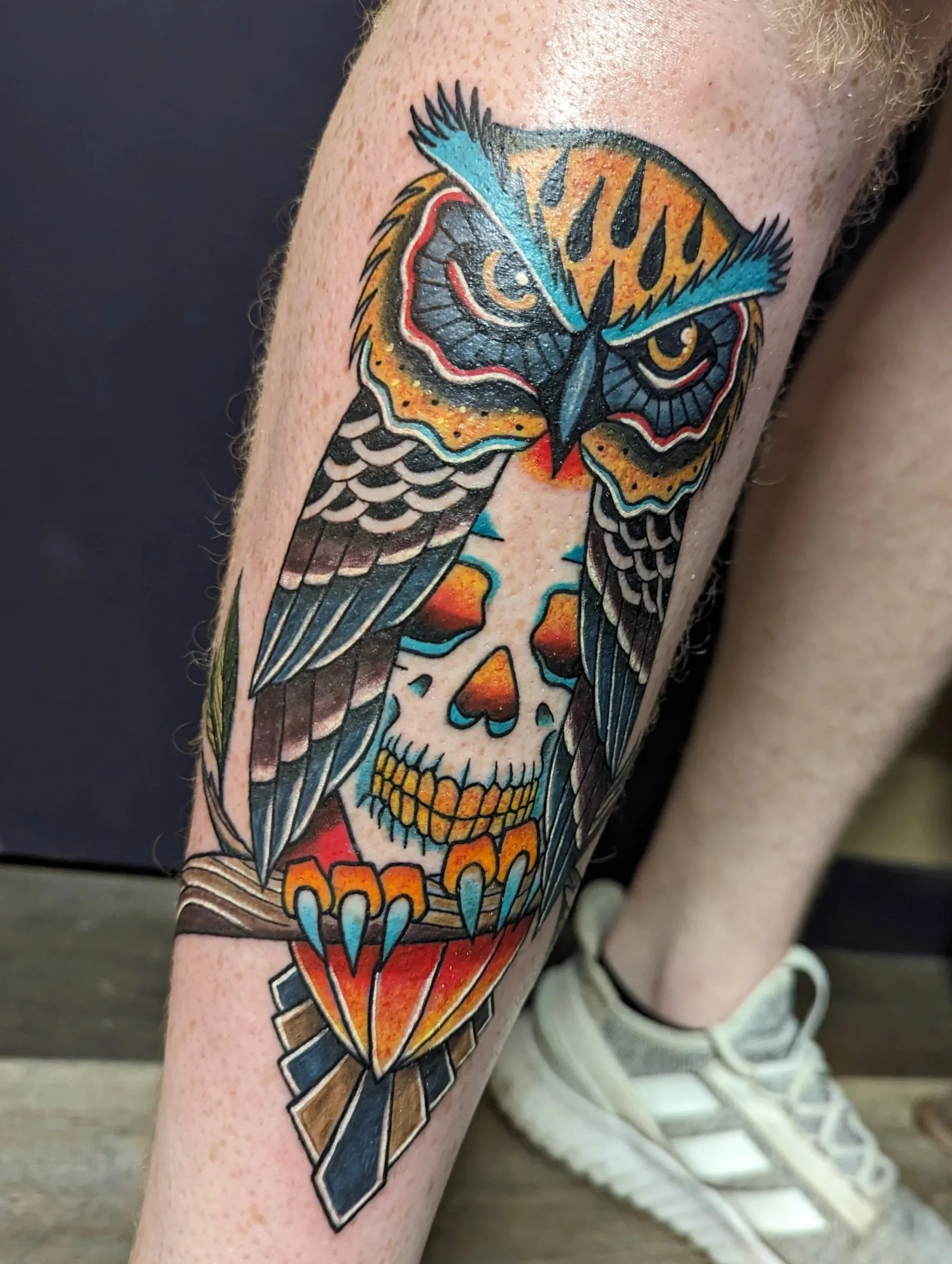 Colorful tattoo of an owl with a skull and bones on a person's leg, with vibrant shades of orange, blue, black, and yellow.