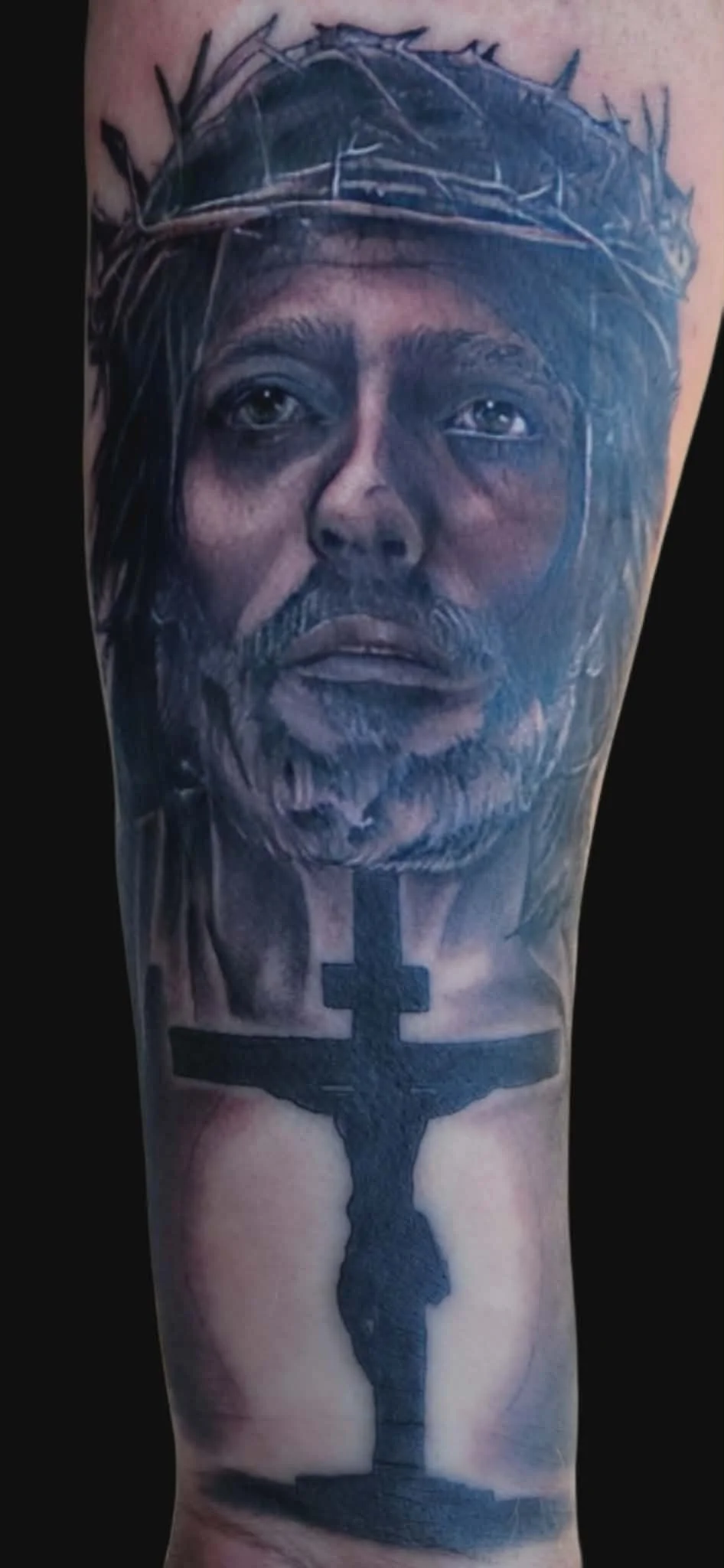 Realistic black and gray tattoo of Jesus Christ's face wearing a crown of thorns, with a crucifix below.