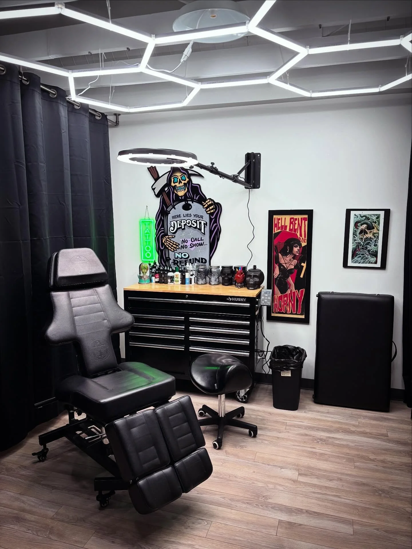 Tattoo studio with black reclining chair, tattoo equipment, and dark art posters, illuminated by modern white ceiling lights.