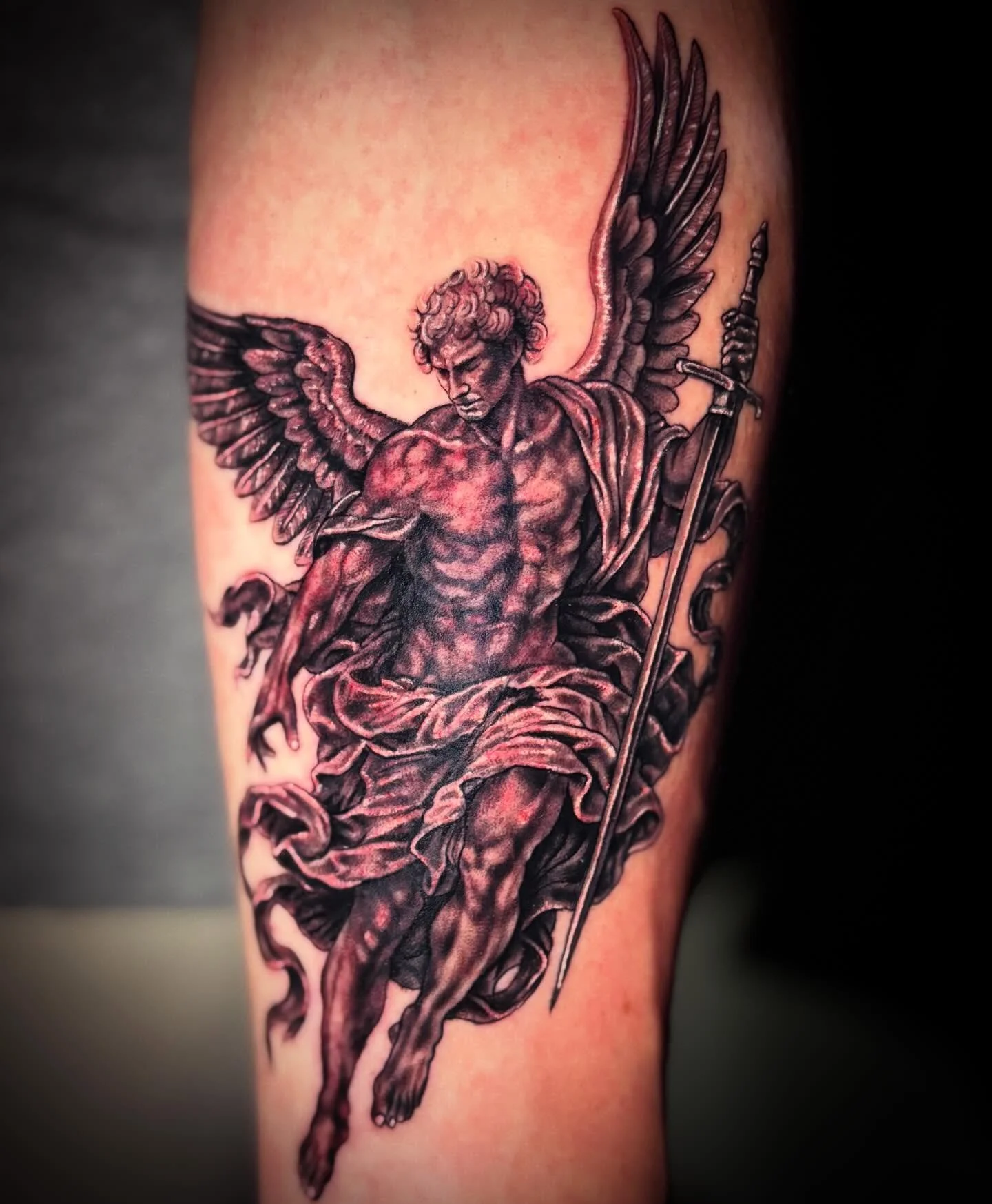 Black and gray tattoo of a muscular angel with large wings, holding a sword, with a serious expression on his face.