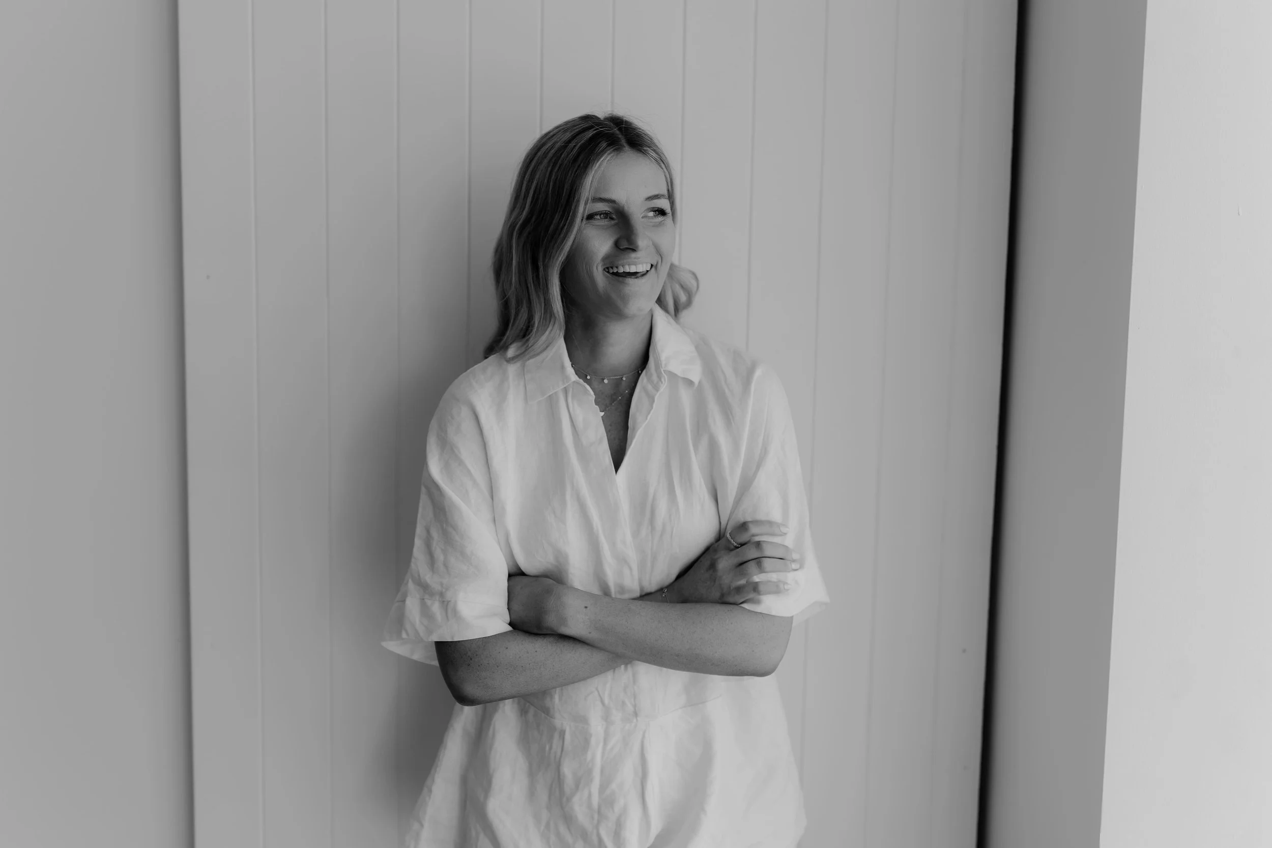 A woman with shoulder-length hair wearing a light-colored shirt, smiling and leaning against a wall with arms crossed.