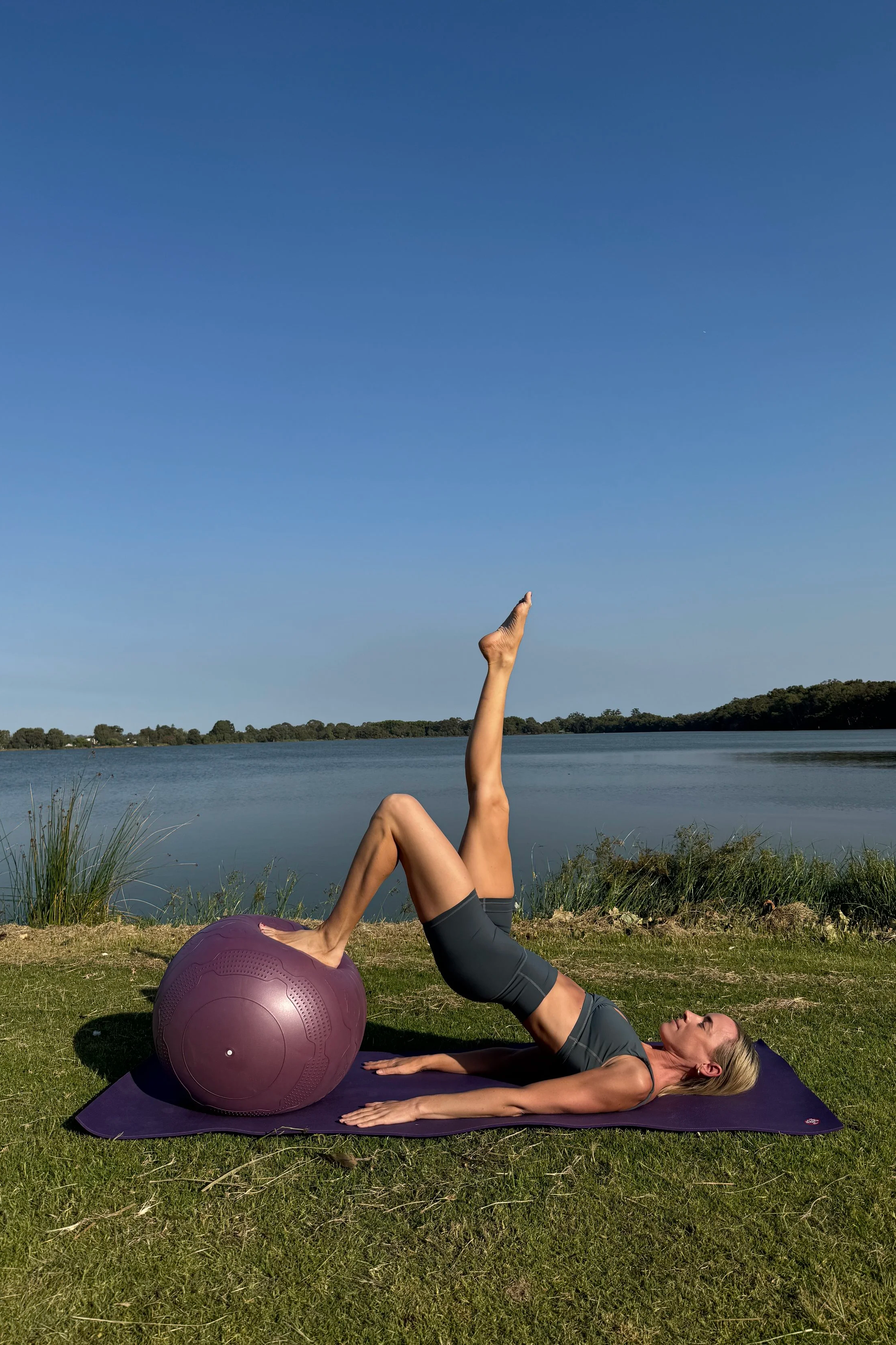 A woman practicing yoga outdoors on a mat next to a lake, with one leg raised vertically and her foot on a large purple exercise ball.