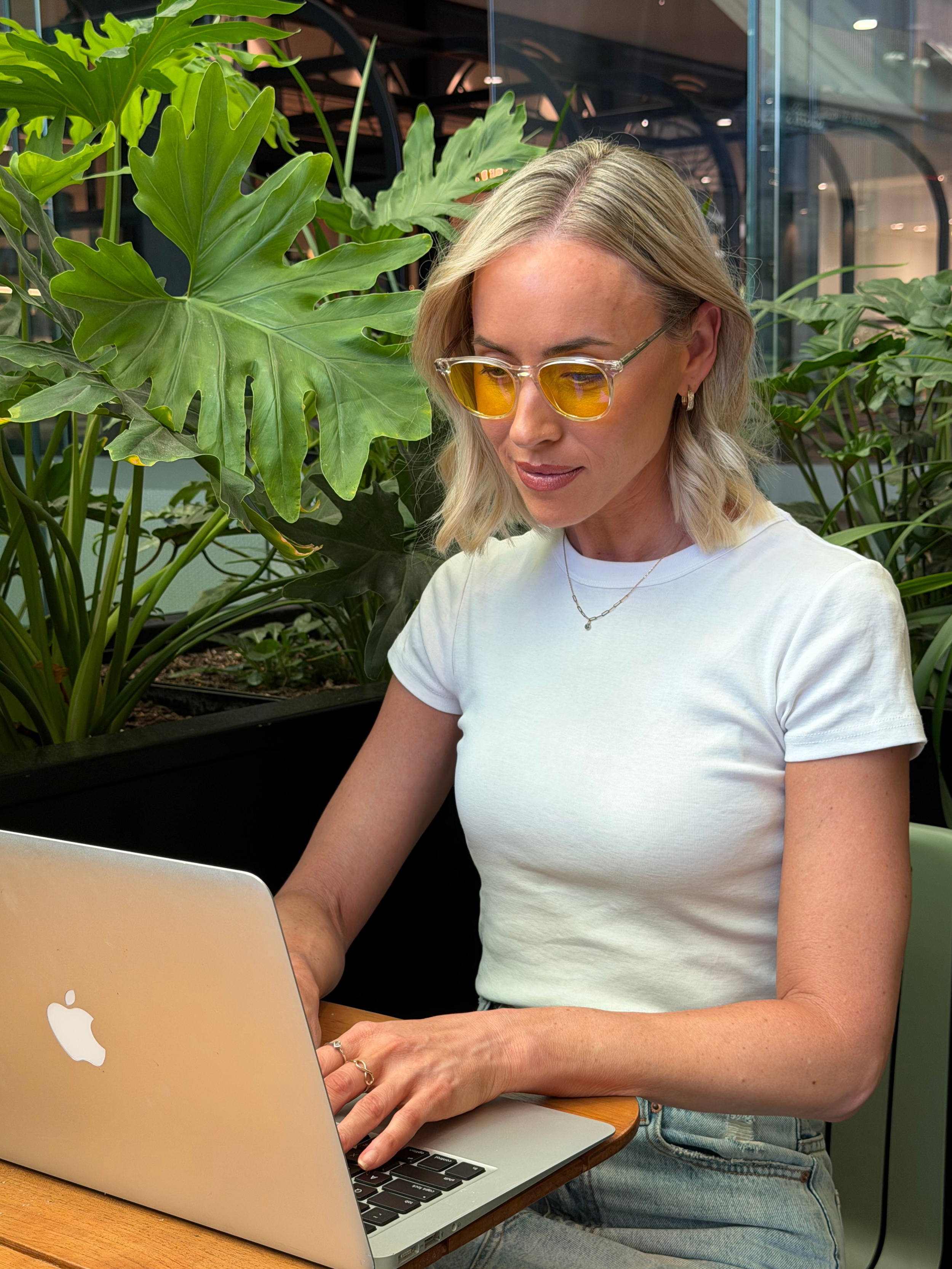 A woman with blonde hair, wearing yellow sunglasses and a white t-shirt, is working on a silver MacBook laptop at a wooden table, surrounded by large green plants in an indoor setting.