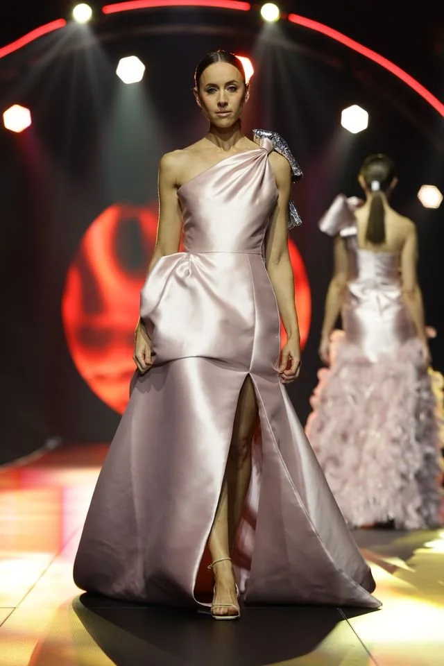 A female model walking on a runway in a shiny, pale pink satin gown with an asymmetric shoulder design and a high slit. The background features red and white lights and another model in a similar gown.