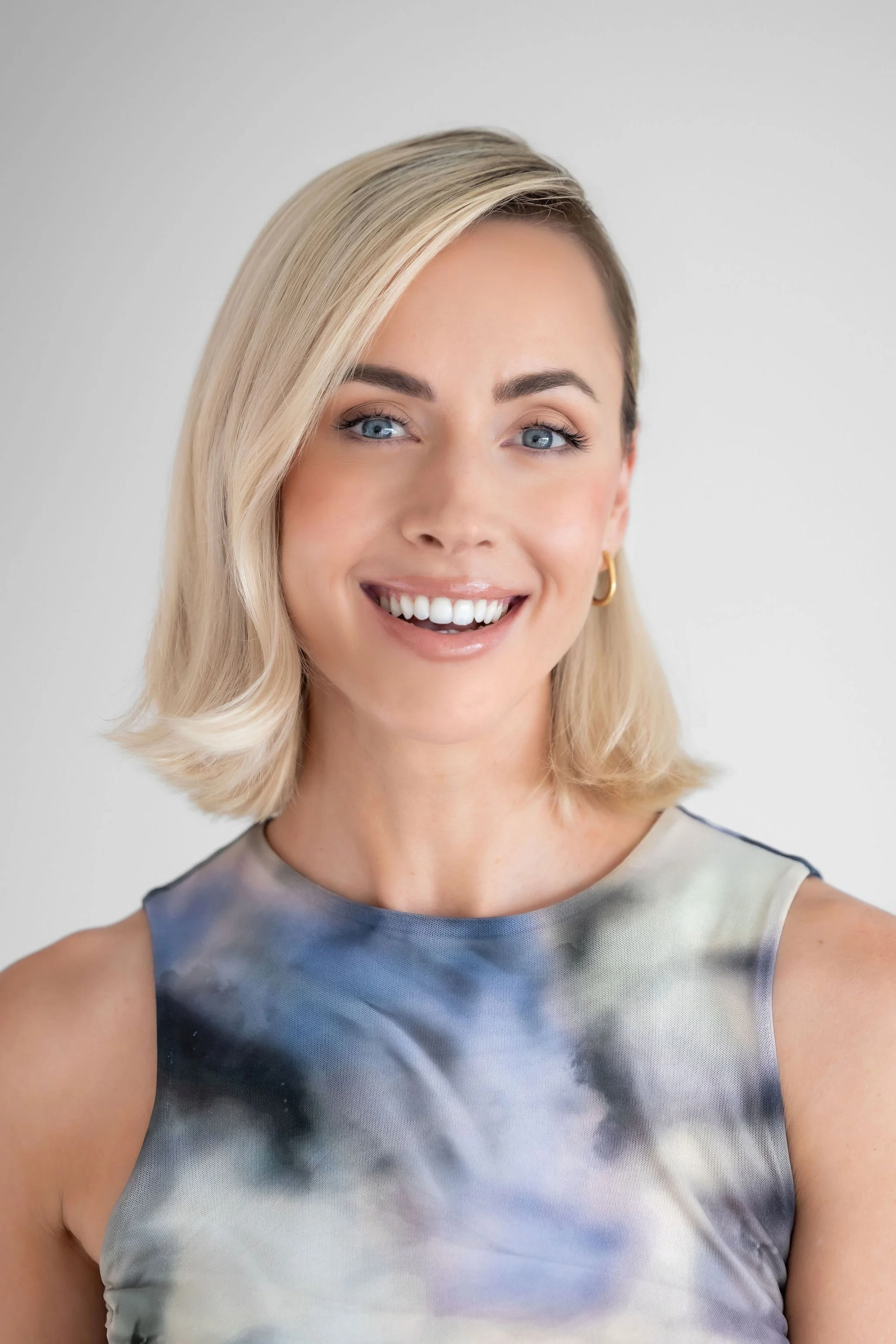 A smiling woman with short blonde hair, blue eyes, wearing a sleeveless dress with an abstract pattern, gold hoop earrings, against a plain white background.