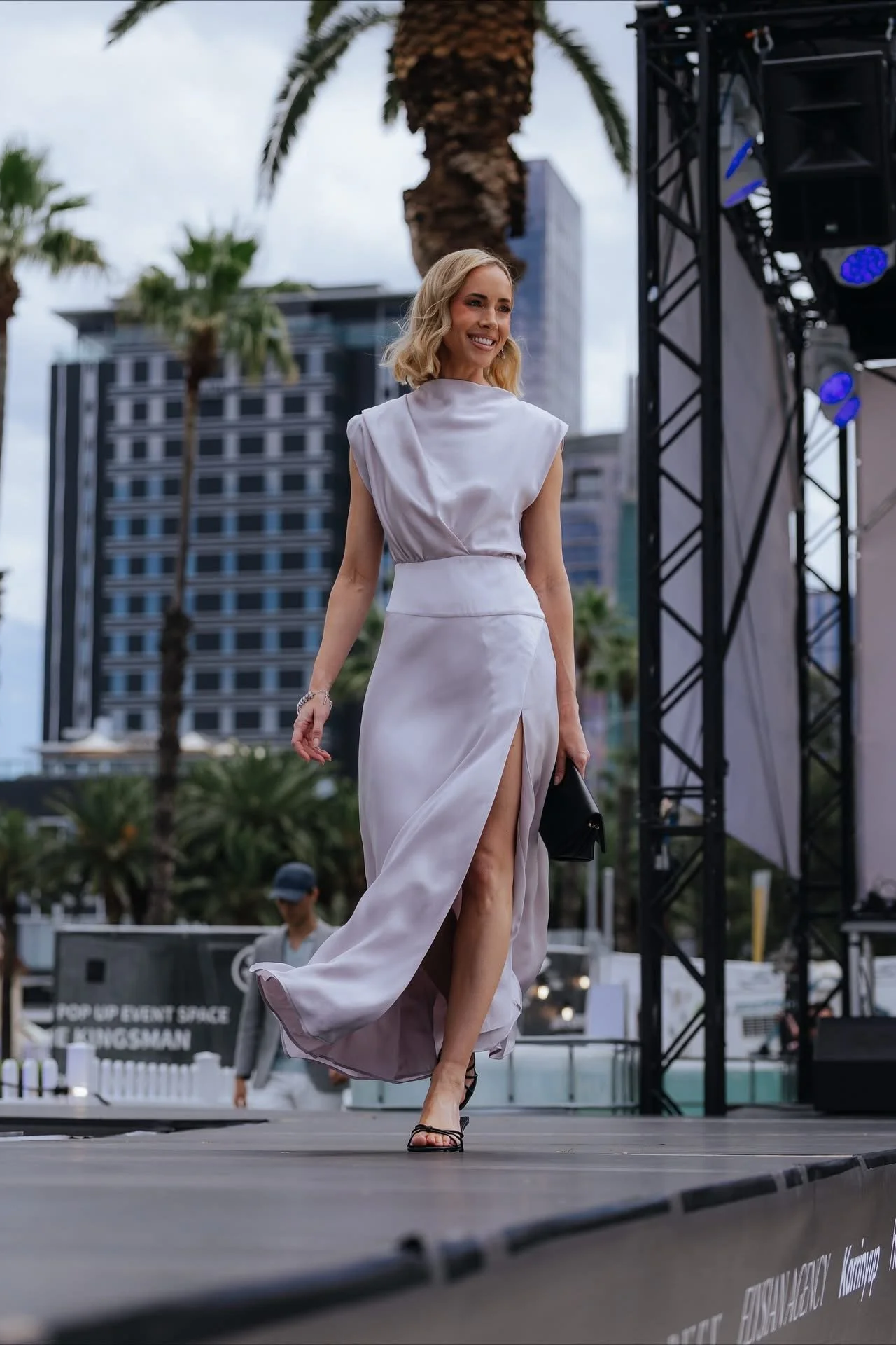 A woman in a flowing, sleeveless, high-slit silver dress walks on an outdoor runway with palm trees and skyscrapers in the background.