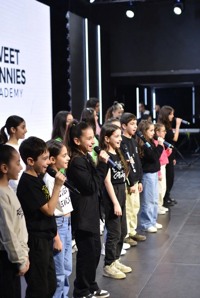 Young children standing on stage holding microphones during a performance at an event, with a screen that has the words 'SWEET BONNIES ACADEMY' in the background.