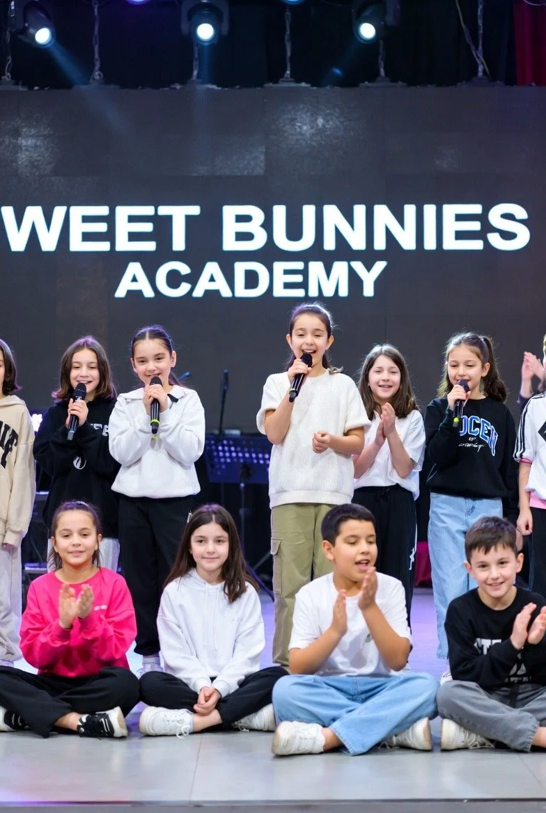 Children on stage during a performance at Weet Bunnies Academy, some holding microphones and others clapping or sitting on the floor, with a large sign behind them displaying the academy's name.