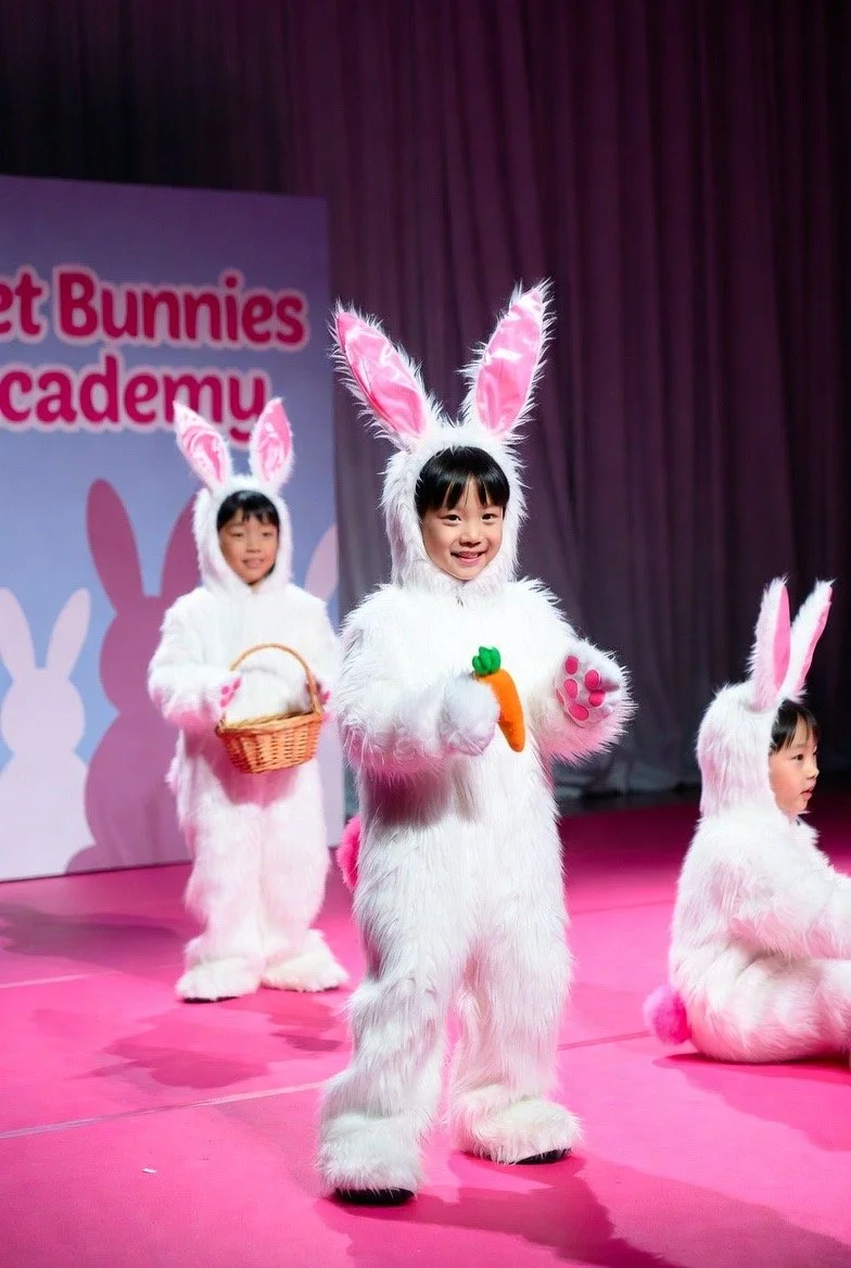 Three children dressed in bunny costumes performing on stage, with one child holding a carrot prop, another with a basket, and the third sitting, in front of a backdrop that reads 'Bunny Bunnies Academy.'