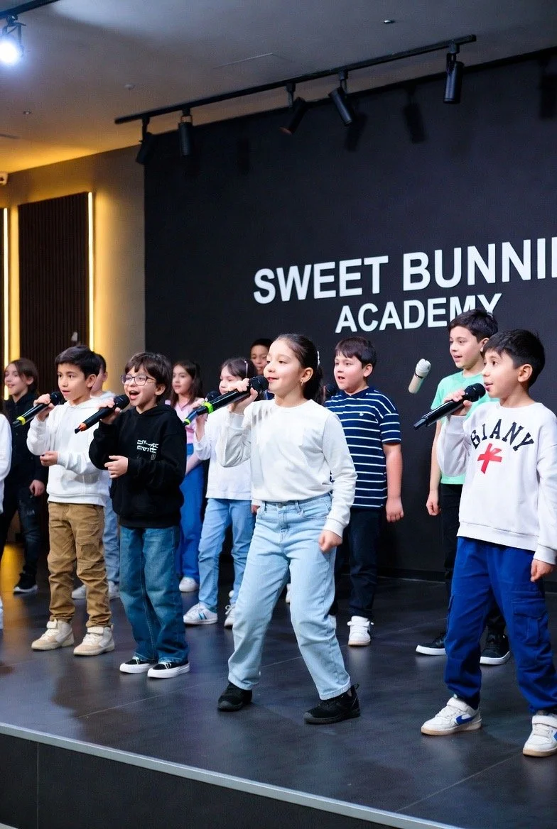 Children singing on stage at Sweet Bunnies Academy, holding microphones.