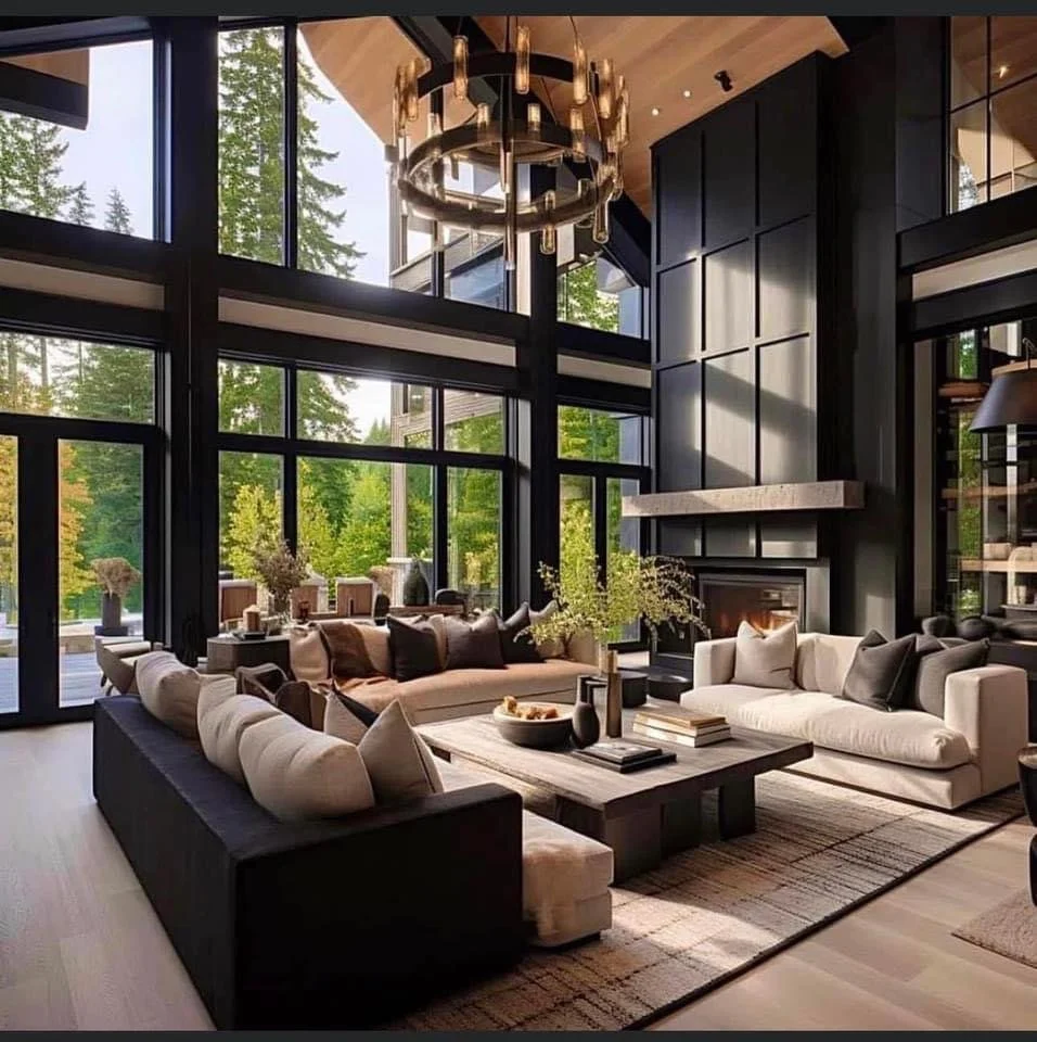 Modern living room with large floor-to-ceiling windows, black framing, a high ceiling, and a circular chandelier. It features a beige sofa, a white sofa, a black sofa, a wooden coffee table with decorative items, and a fireplace with a black wall behind it.