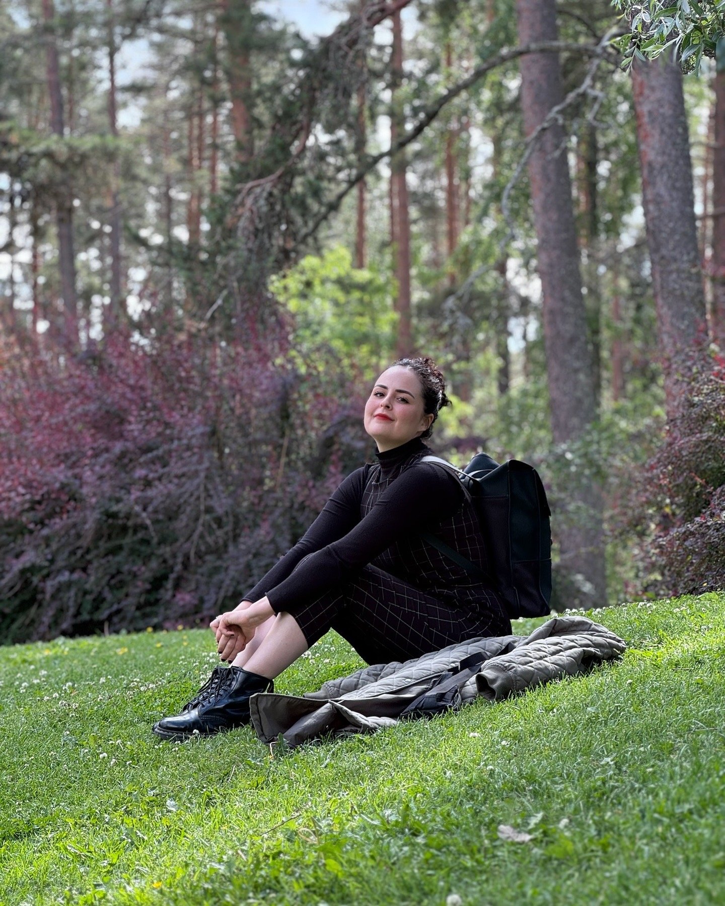 Persona sentada en un parque, rodeada de árboles y vegetación, con una chaqueta verde y mochila negra.