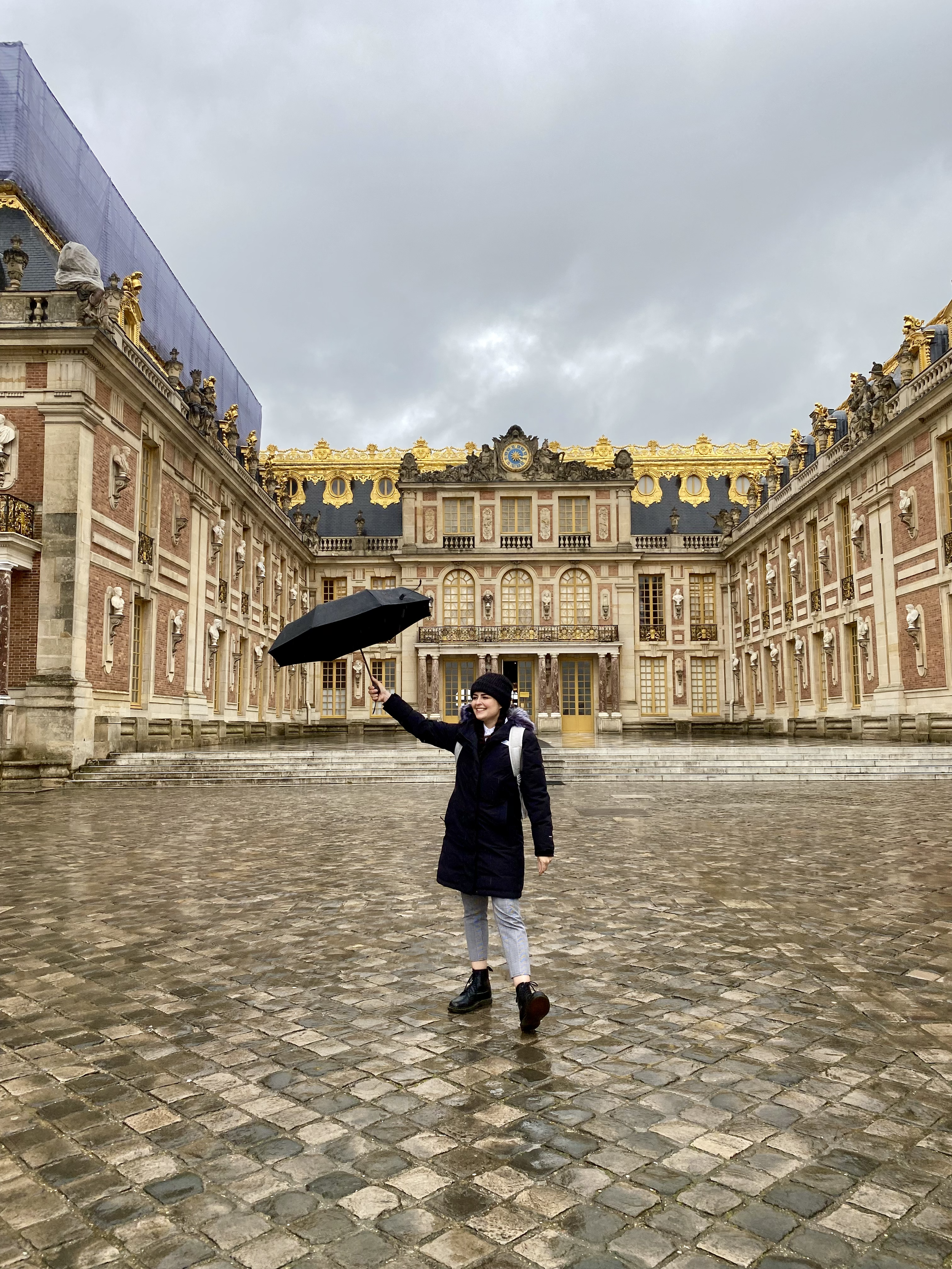 Persona con paraguas negro frente a un edificio histórico con adornos dorados y arquitectura clásica, cielo nublado.