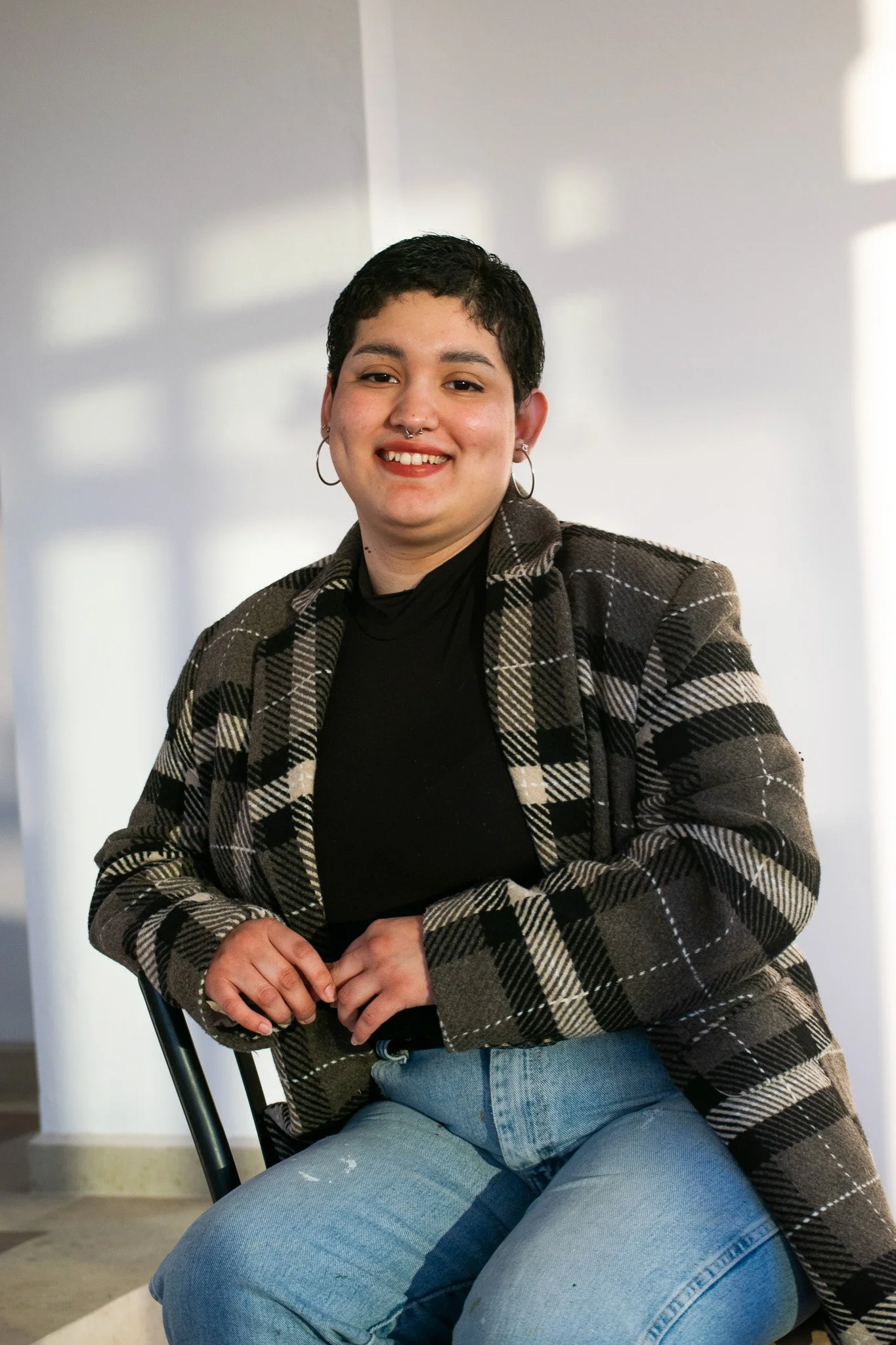 Persona sonriendo, con cabello corto, usando un abrigo a cuadros, camiseta negra y jeans, sentada en una silla.