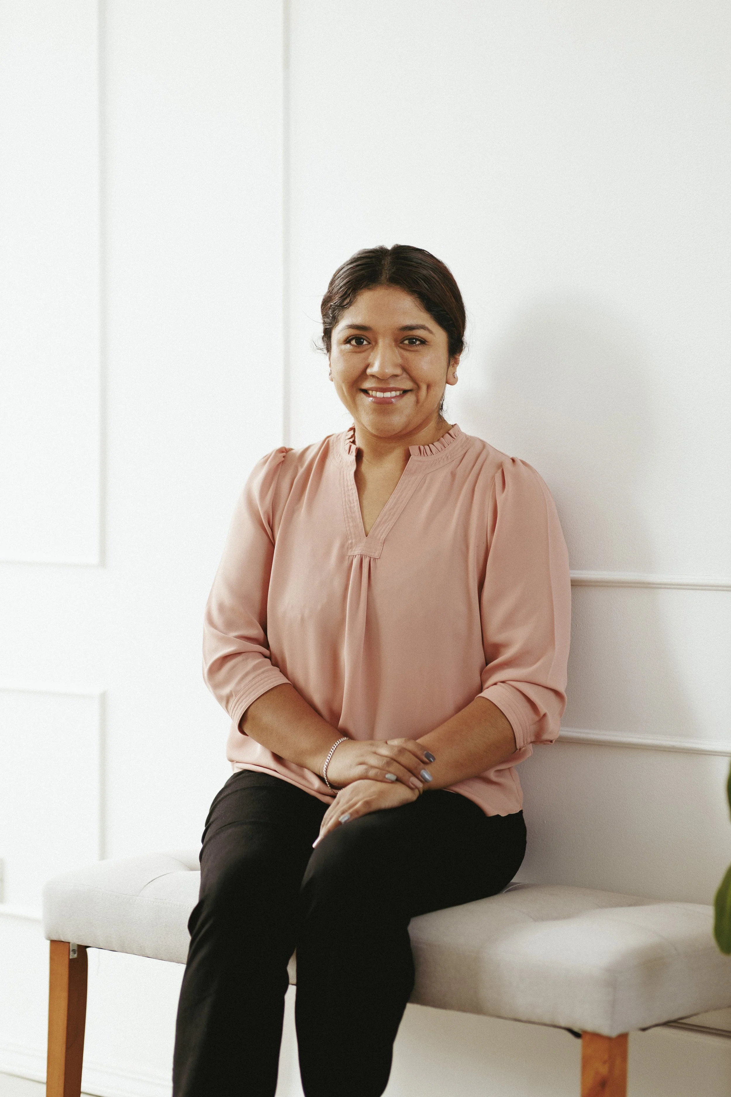 Persona sentada en un banco, vestida con blusa rosa y pantalones negros, sonriendo frente a una pared blanca.