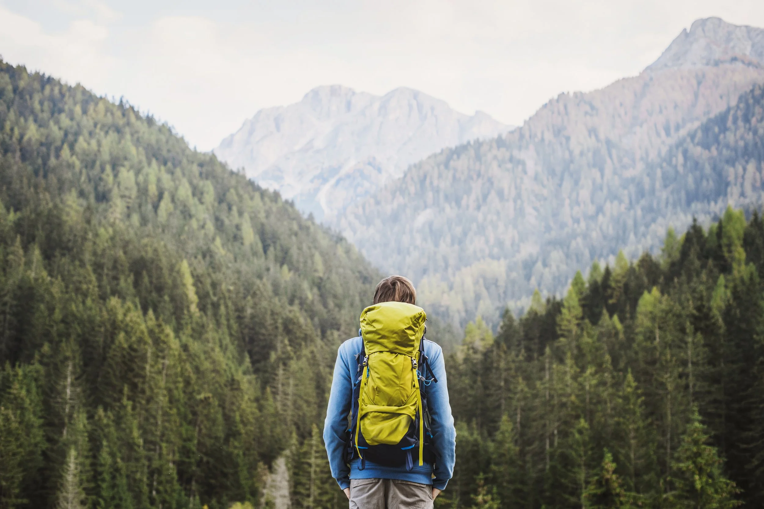 Persona con mochila amarilla, de pie frente a un paisaje montañoso y boscoso.