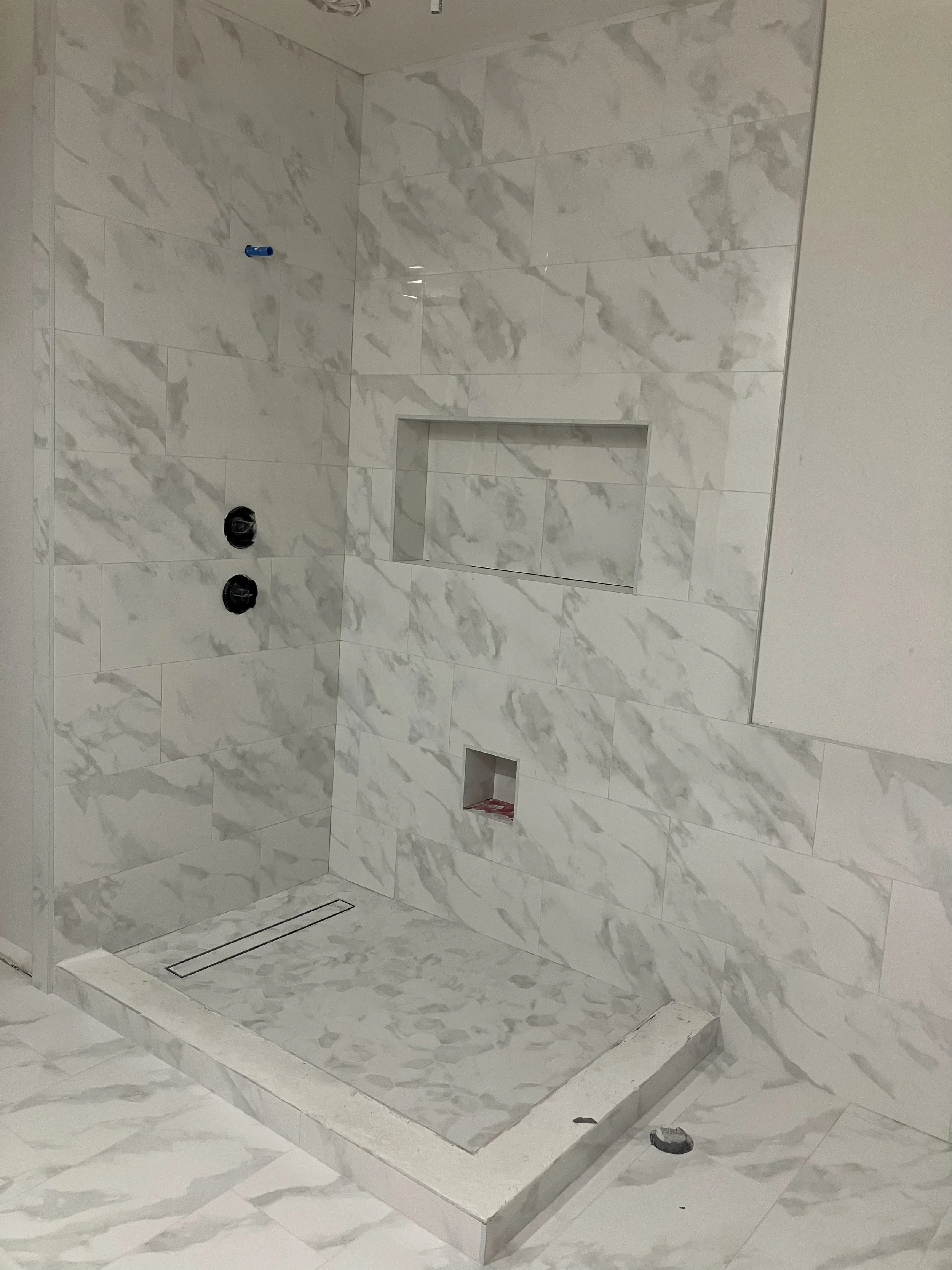 A partially completed walk-in shower with marble tiles, a built-in niche, and plumbing fixtures without a shower door or curtain.
