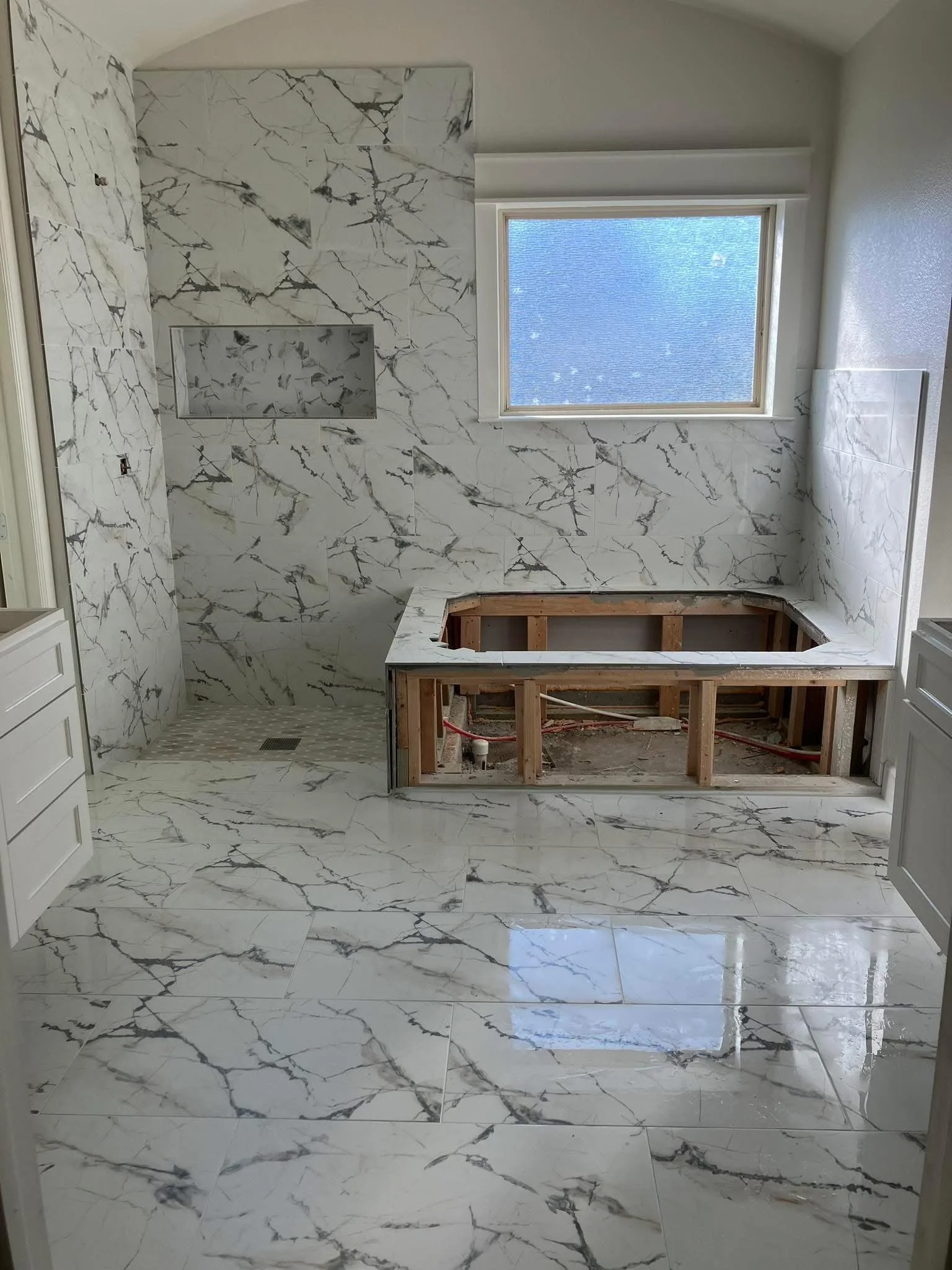 Bathroom under construction with marble tile walls and floor, a window, and an open framed bathtub area.