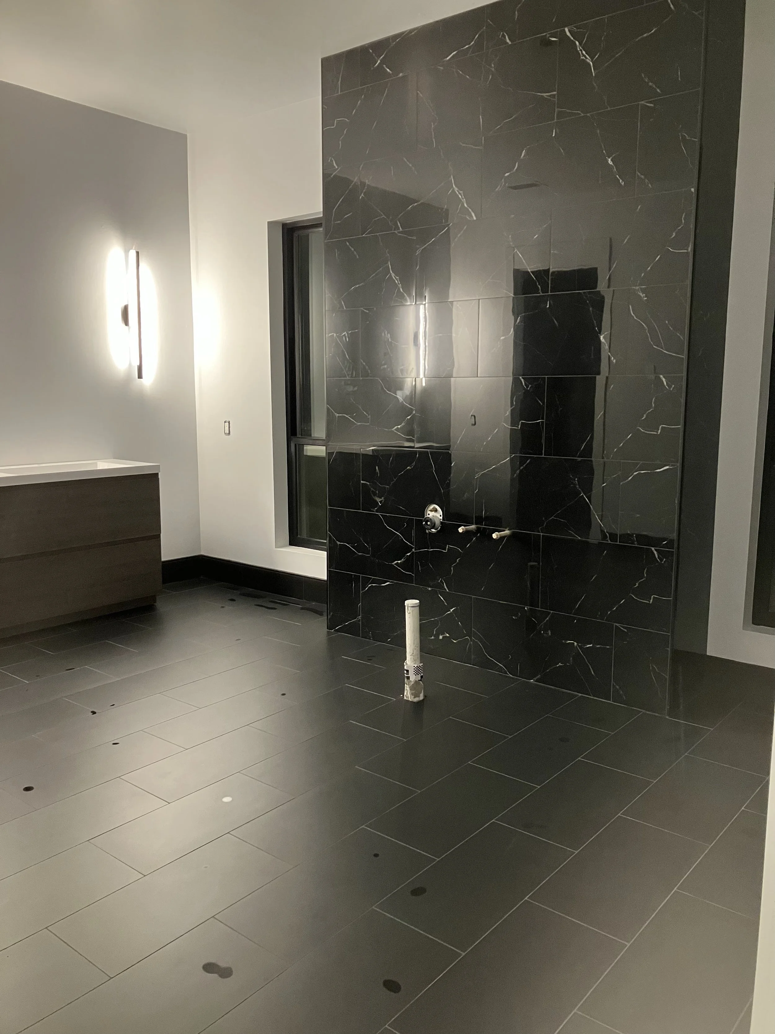 Interior of a modern bathroom or spa area with gray tiled walls and floor, a black marble accent wall, a window, and a wall-mounted light fixture.