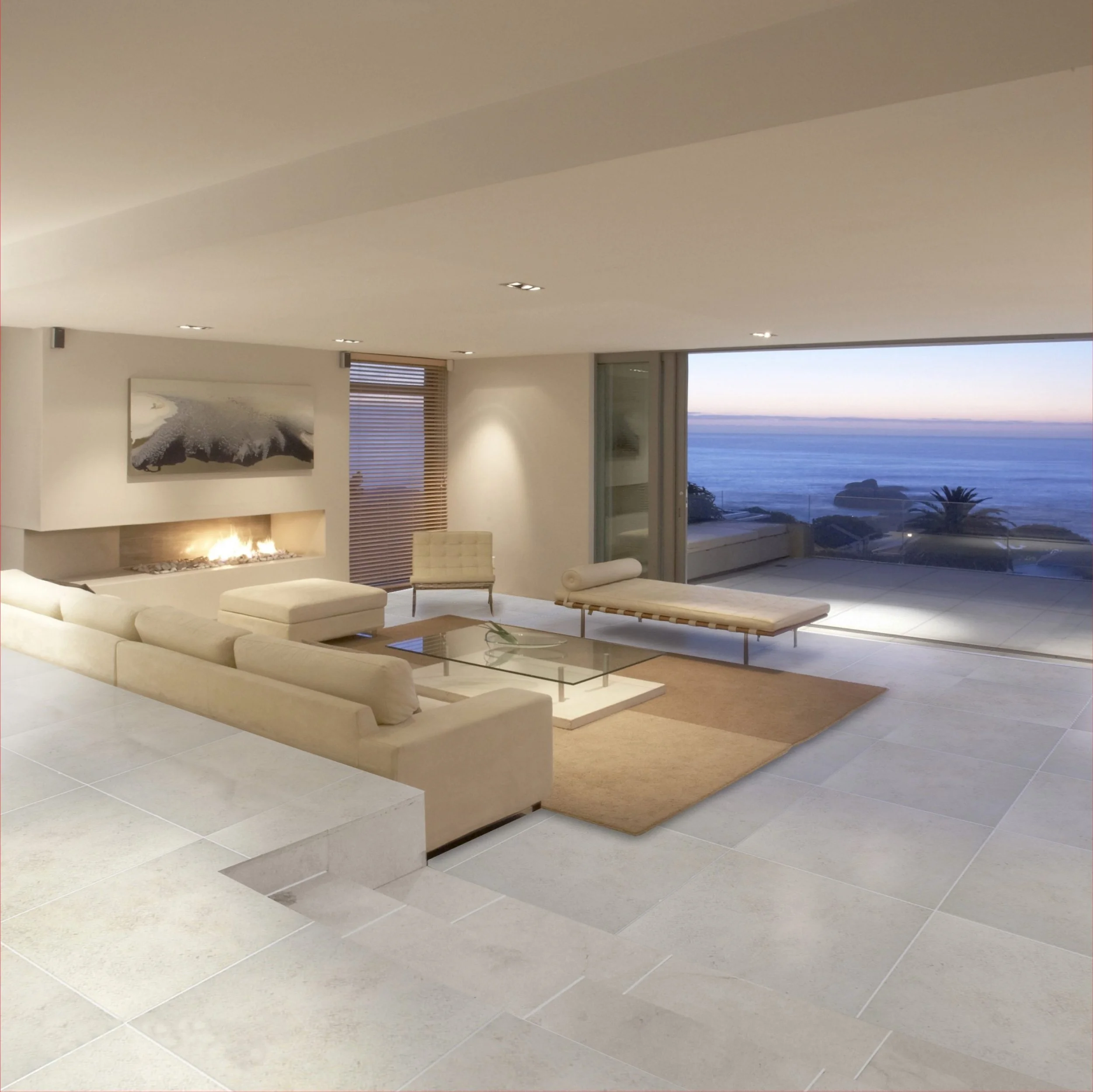 Interior of a modern, minimalistic living room with beige furniture, a fireplace, artwork on the wall, large windows with blinds, and an ocean view outside.