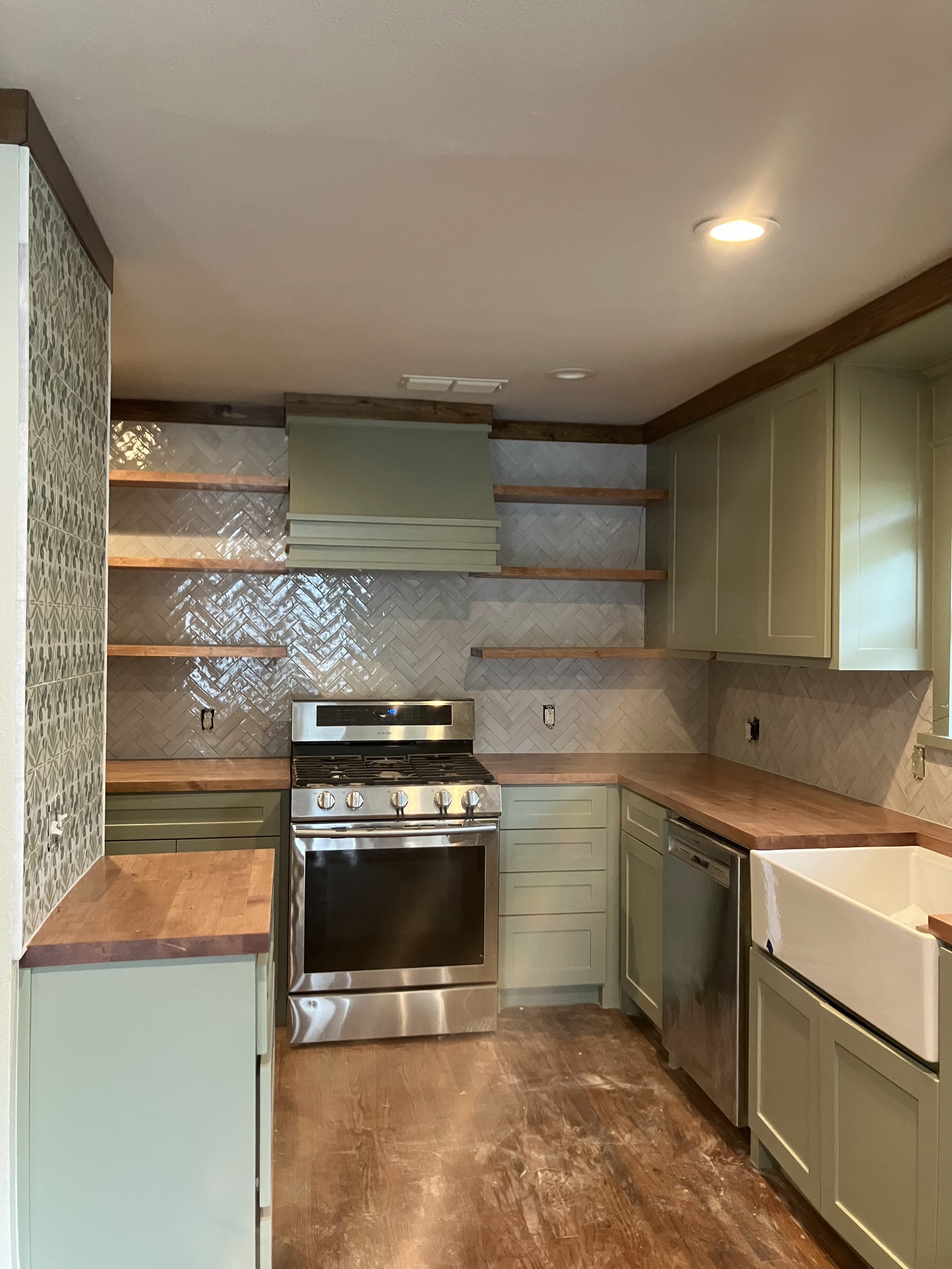 A kitchen with light green cabinets, wooden shelves, a gray tile backsplash, a stainless steel oven, and a beige ceiling with recessed lighting.