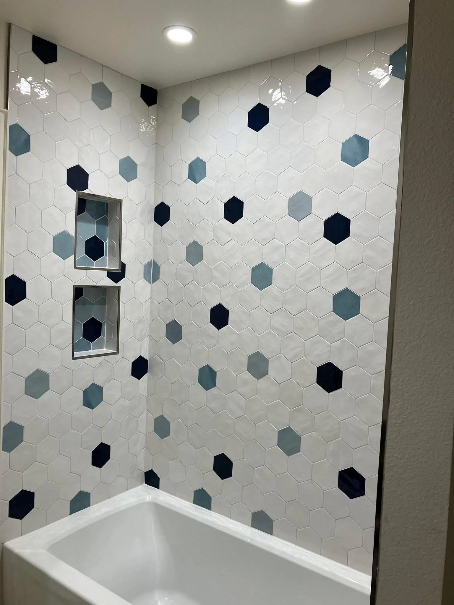 Bathroom wall with white hexagonal tiles, some tiles are dark blue, gray, and black. There are two built-in wall niches with matching hex tile interiors. A white bathtub is visible at the bottom of the image, and a ceiling light is above.