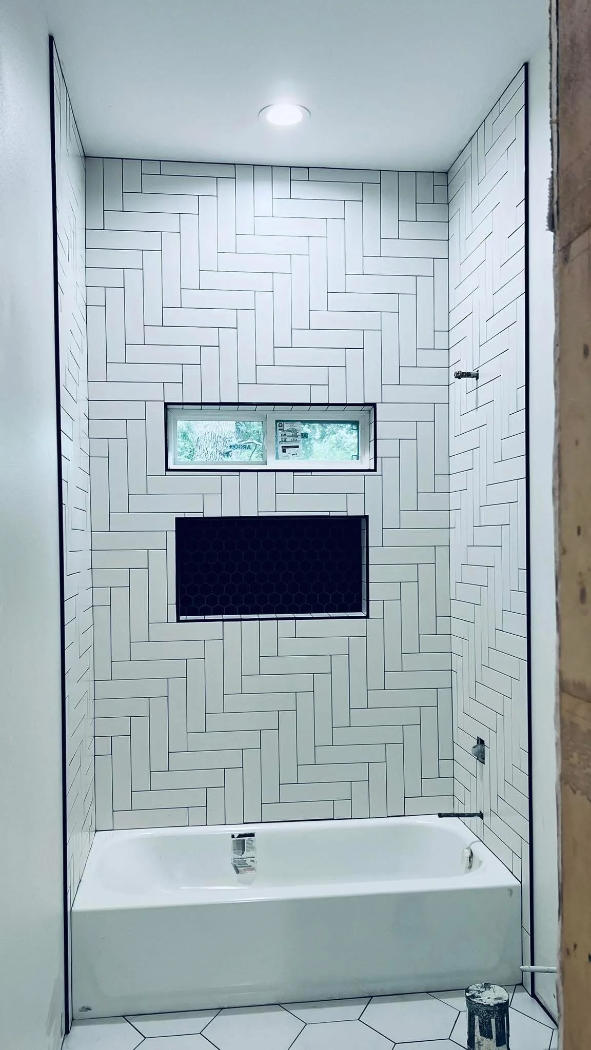 A bathroom with a white ceramic bathtub, white hexagonal floor tiles, and a wall with white rectangular tiles arranged in a herringbone pattern. There is a small rectangular window with a view of trees outside and some black hexagonal tiles behind a niche. An overhead light illuminates the space.