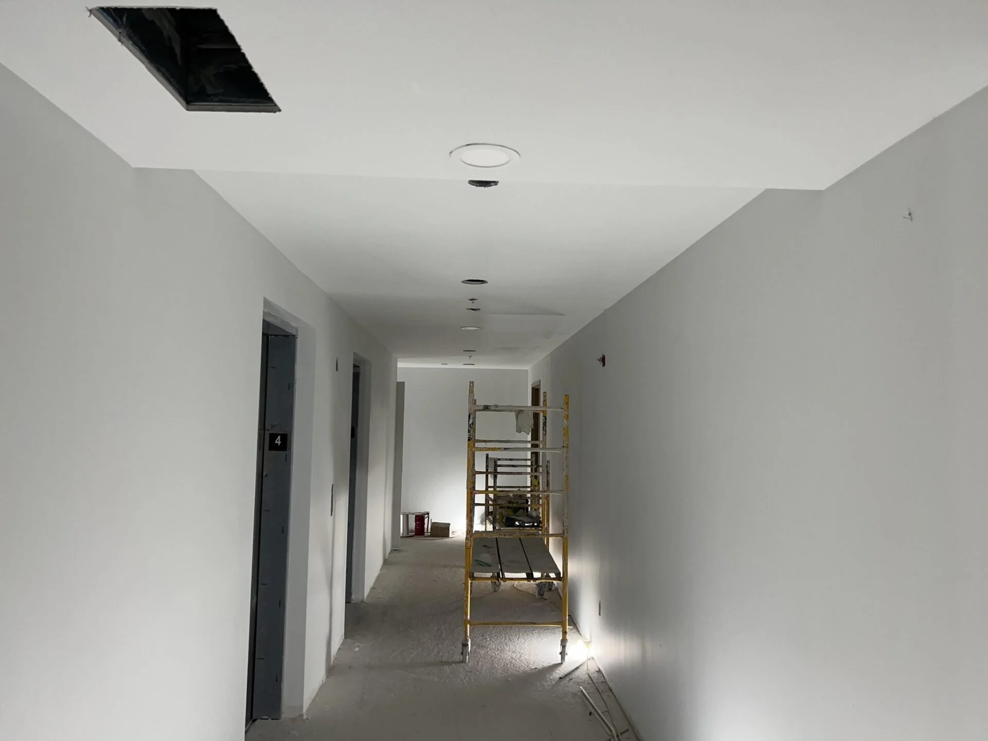 Long hallway in a building under construction or renovation, with white walls, open elevator doors on the left, scaffolding and construction materials in the background, and exposed ceiling with lighting fixtures.