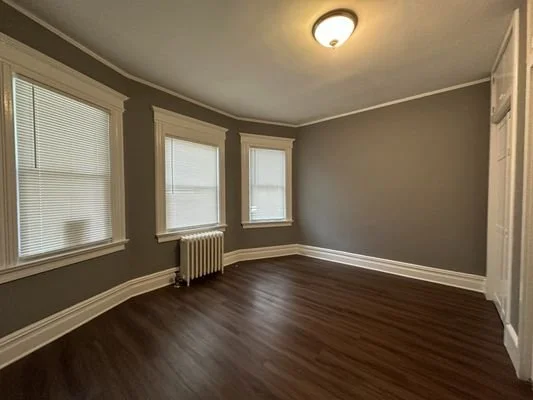 Empty room with three windows, a radiator, hardwood floors, and a ceiling light fixture.