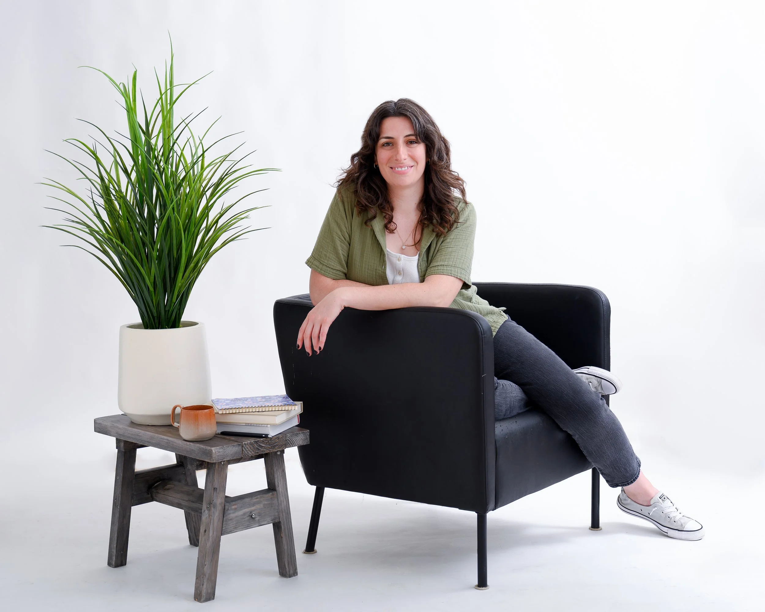 Young woman with curly hair smiling while sitting on a black armchair, with her feet up on the chair. There is a large green plant in a white pot on a small gray wooden table next to her, with notebooks, a small ceramic cup, and a notebook on the table. The background is plain white.