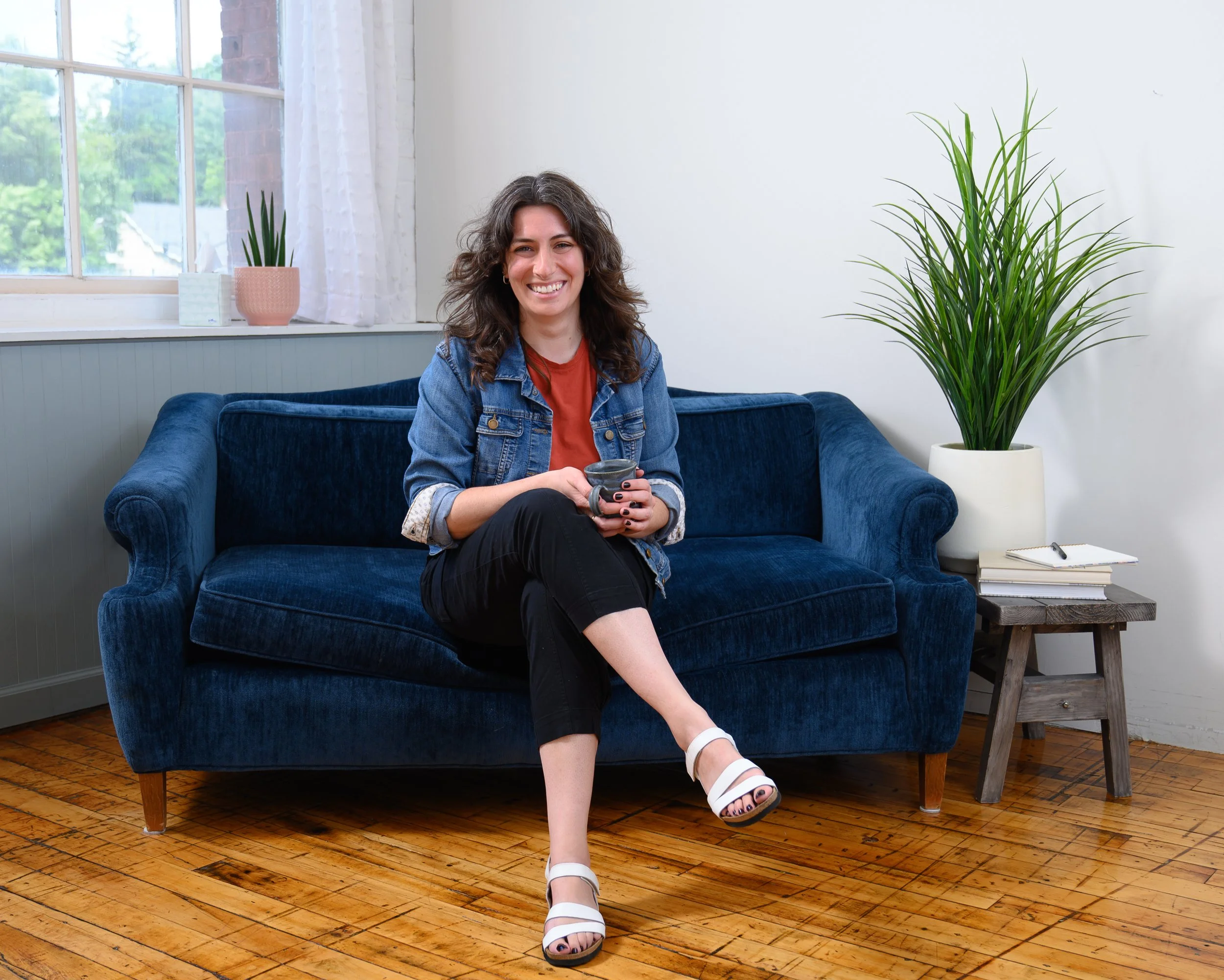 A woman with curly brown hair, wearing a denim jacket and black pants, is sitting on a blue velvet sofa in a bright room. She is smiling and holding a mug. There is a large green plant in a white pot on a small wooden side table next to her, with some books and a notebook. A window with white curtains is in the background, showing outside greenery.
