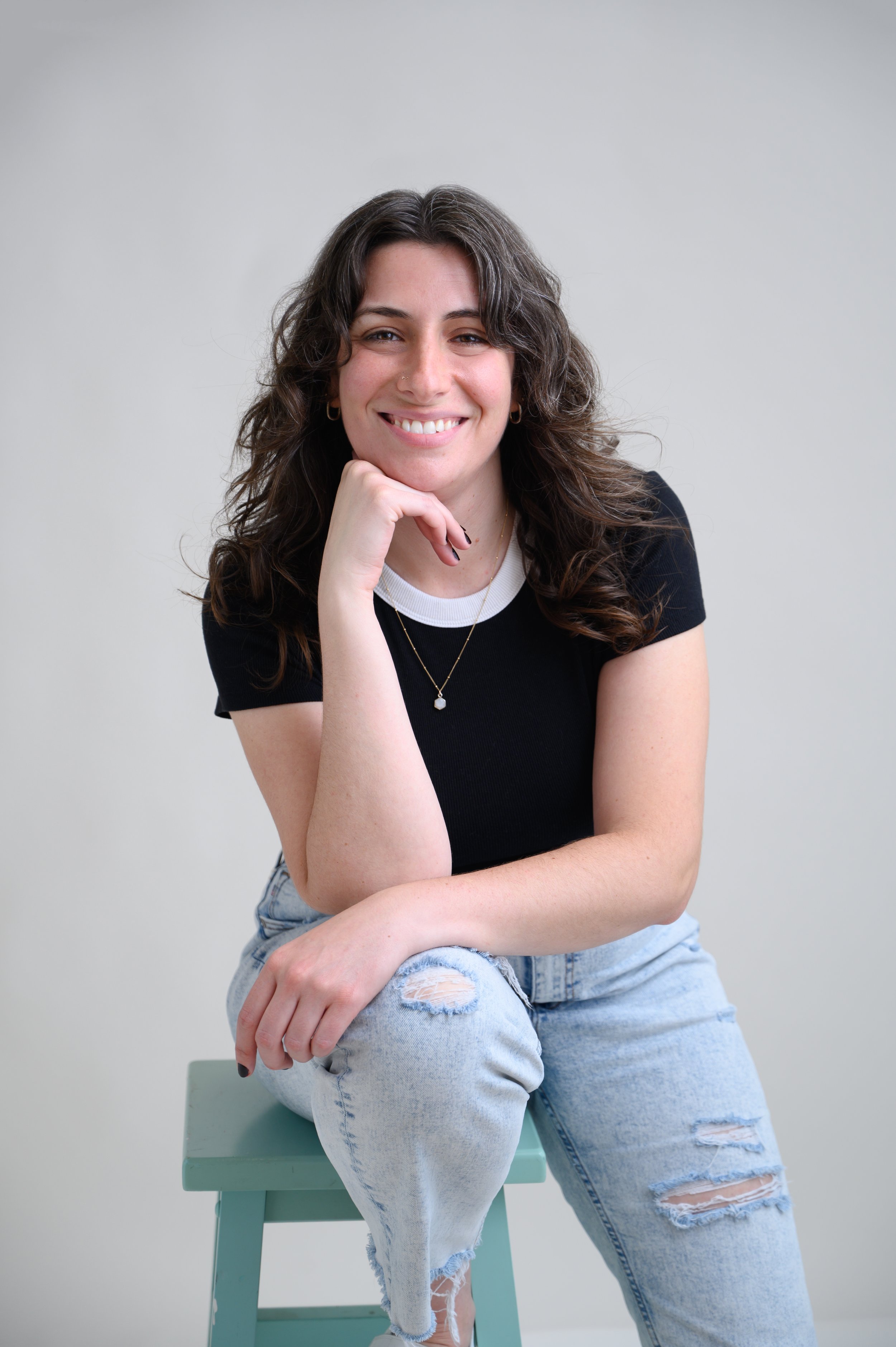 A young woman with wavy brown hair smiling, sitting on a light green stool, leaning her chin on her hand, wearing a black t-shirt with white collar, distressed light blue jeans, and jewelry, in front of a plain gray background.