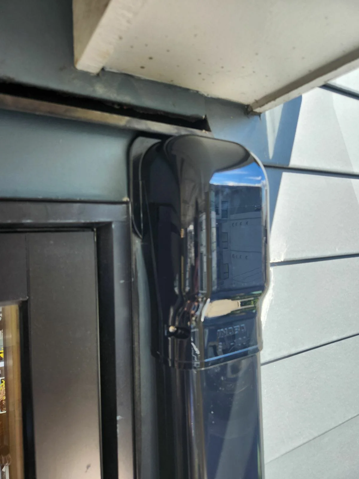 Close-up of a black coffee machine on a countertop outside, with a house and blue sky reflected in its shiny surface.