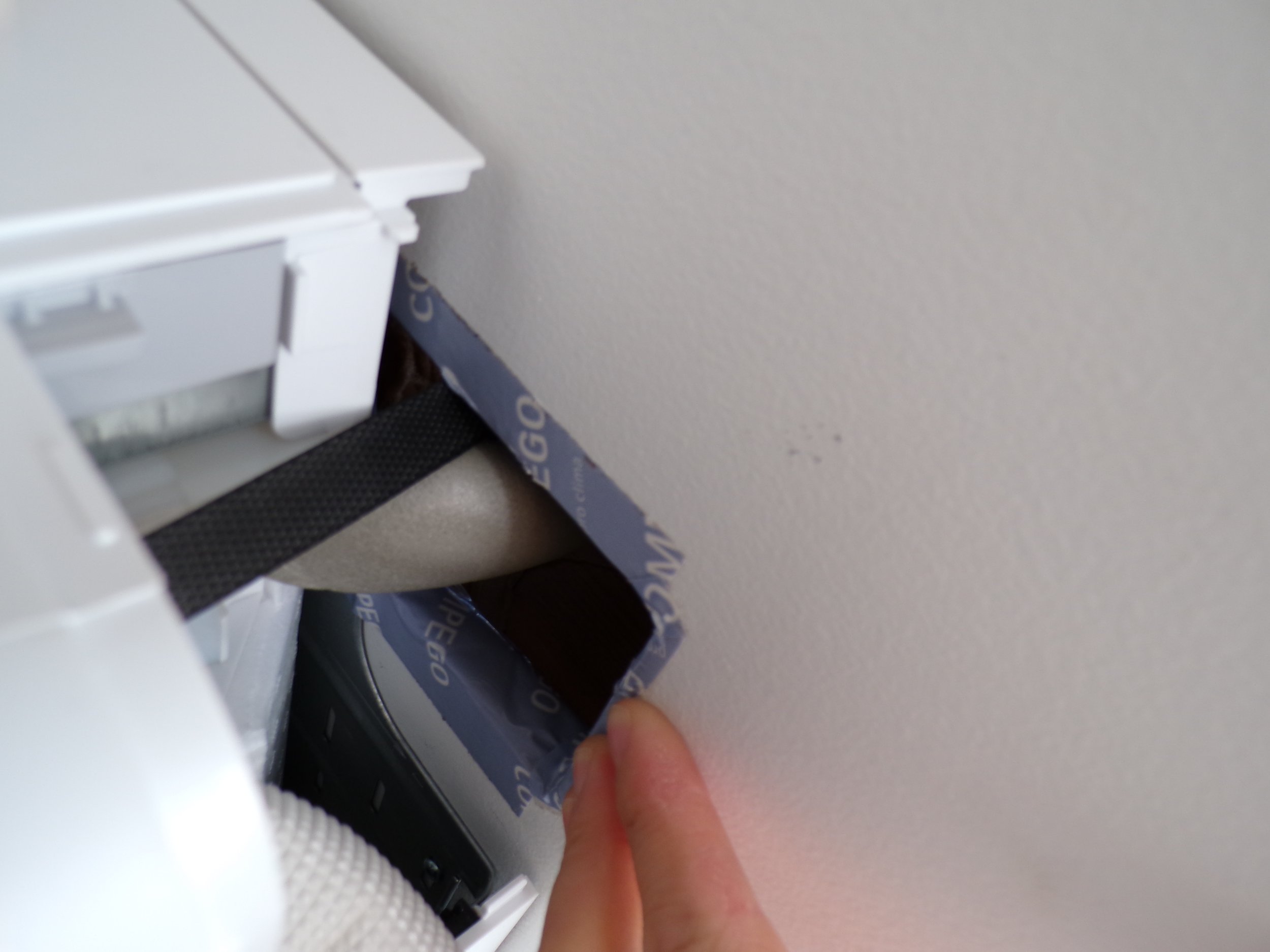 A close-up of a person's hand peeling back a piece of blue adhesive tape from a wall socket or outlet cover.