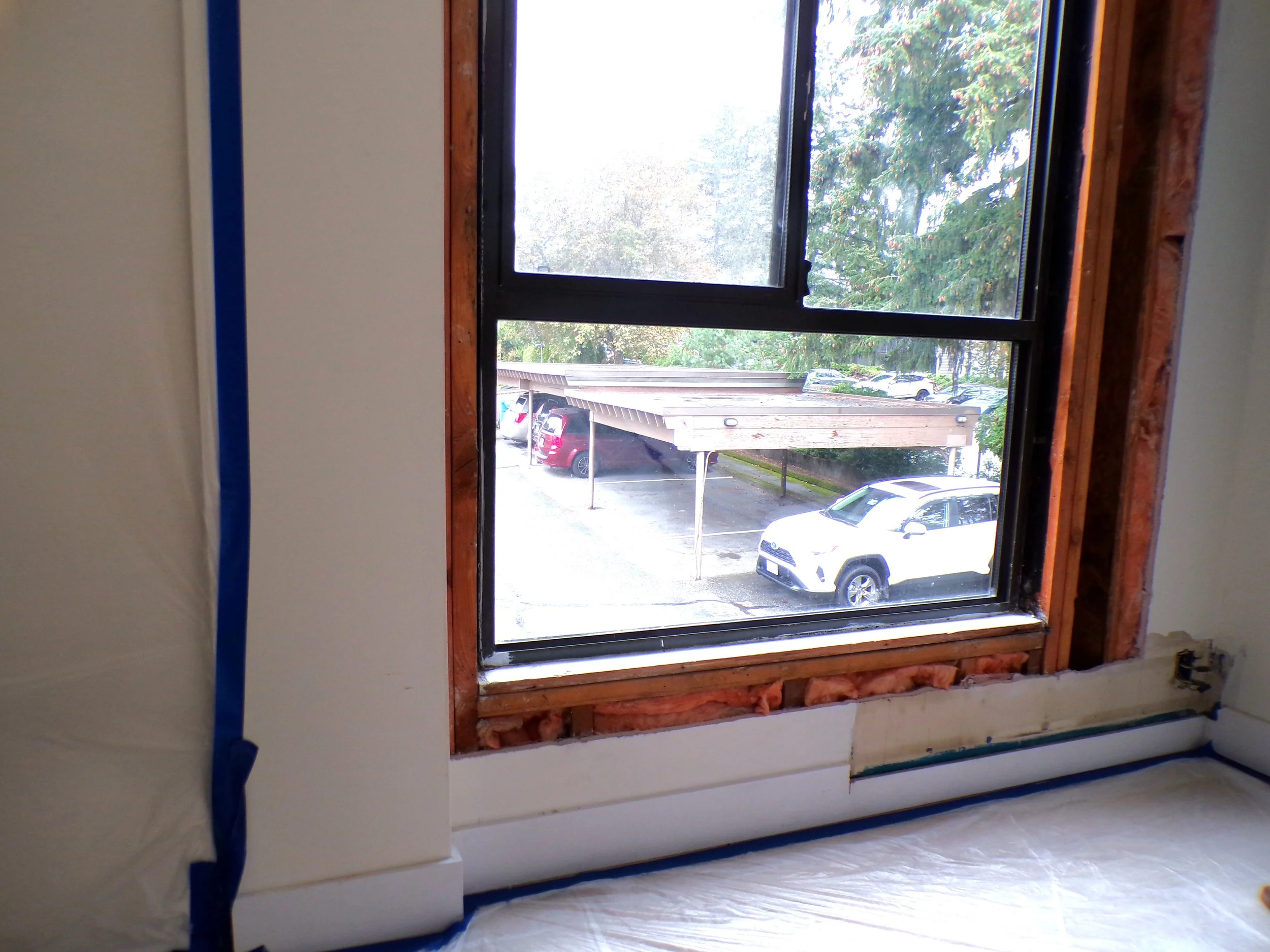 Interior view of a room under renovation or construction, showing a window with exposed framing and insulation around the edges, with blue painter's tape along the bottom and left sides, and a white protective sheet on the floor.