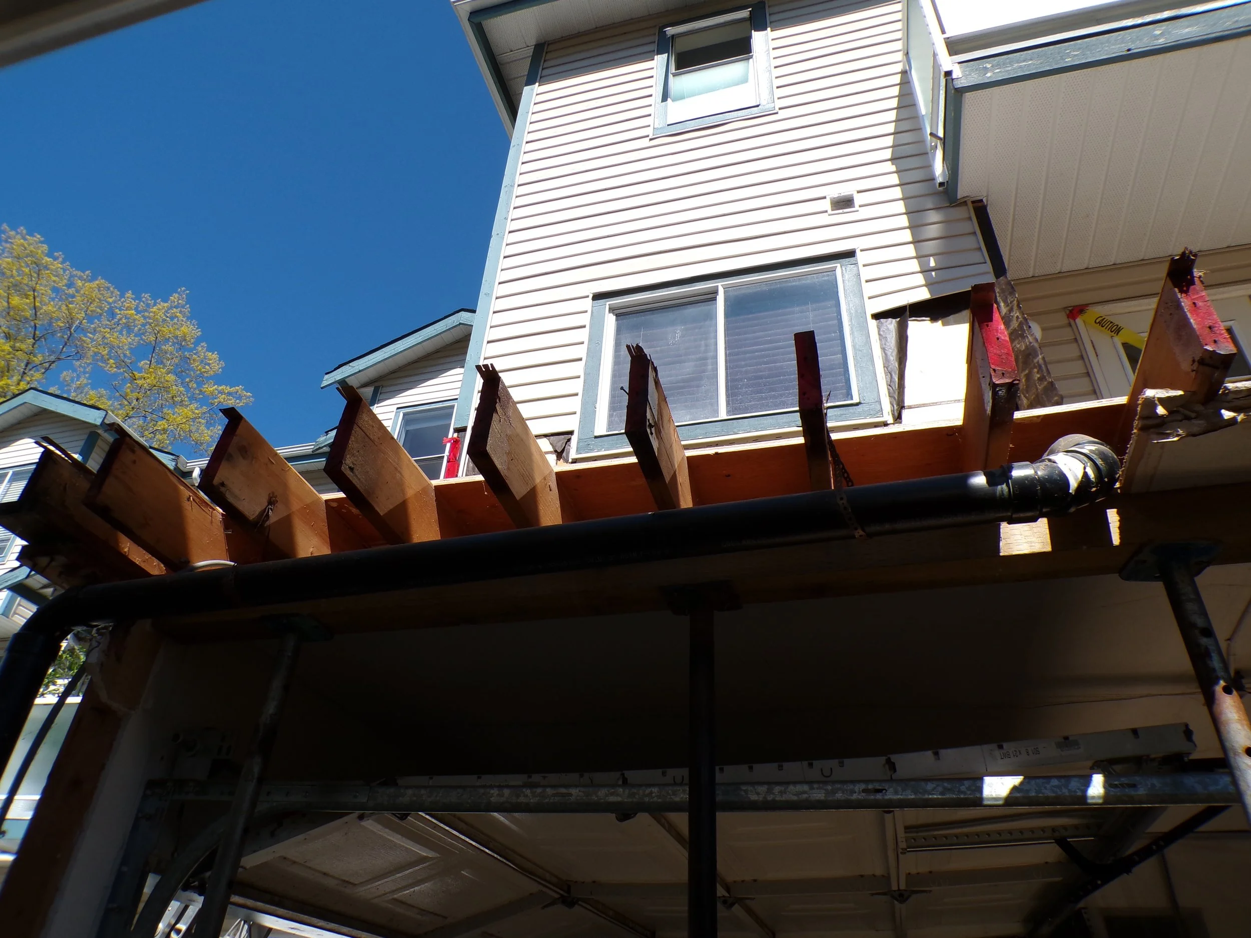 Construction work on a house with a focus on the edge of the second story, showing exposed wooden beams, a black drainage pipe, and part of the balcony, under a clear blue sky.