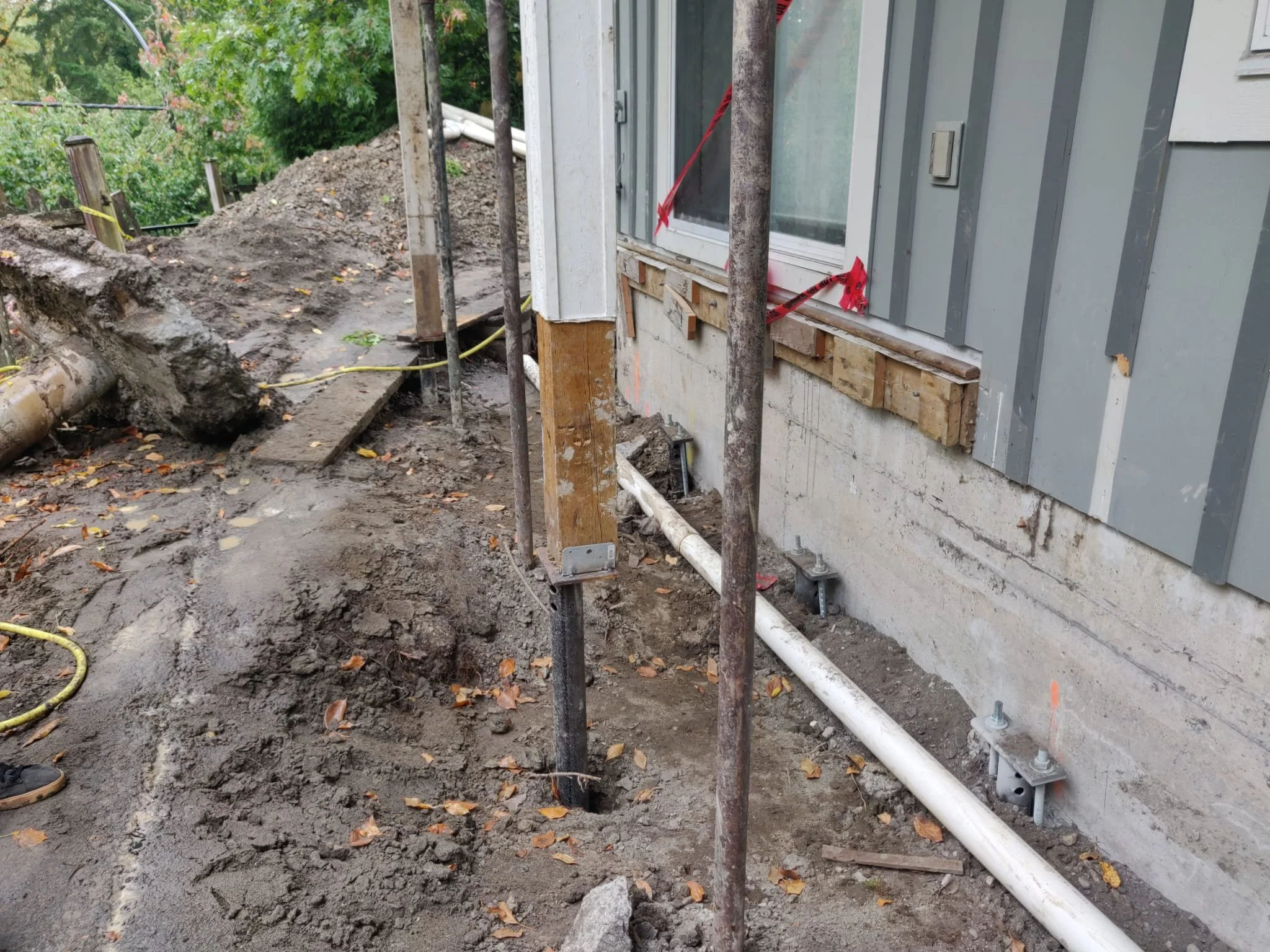 Construction site with scaffolding and support beams along the side of a house, with exposed foundation and pipes visible, and dirt and leaves on the ground.